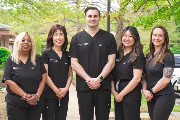 Five staff members in black uniforms standing outdoors in front of greenery and a building.