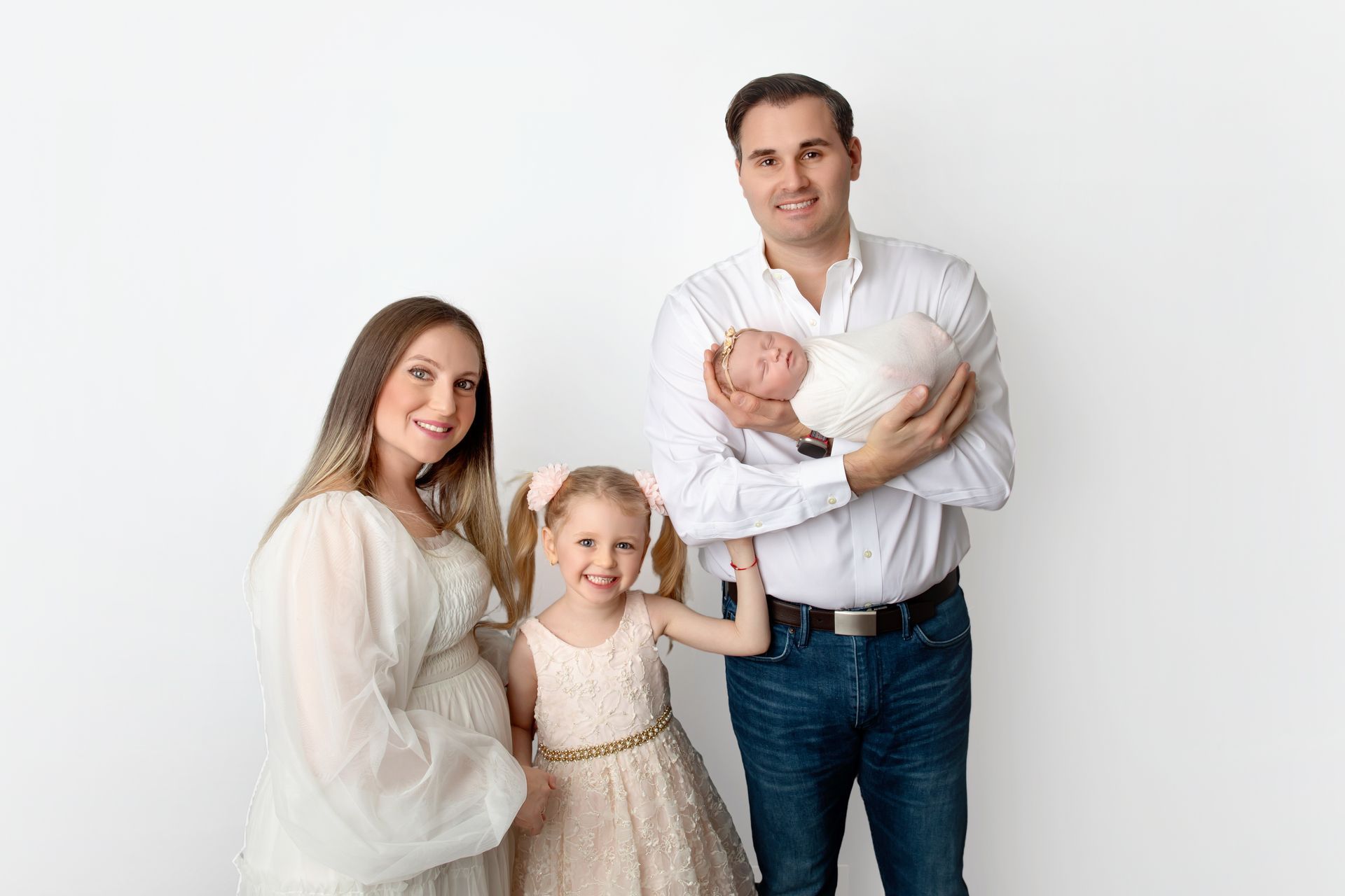 Family of four posing against a white background, with parents holding a newborn and a young child standing beside them.