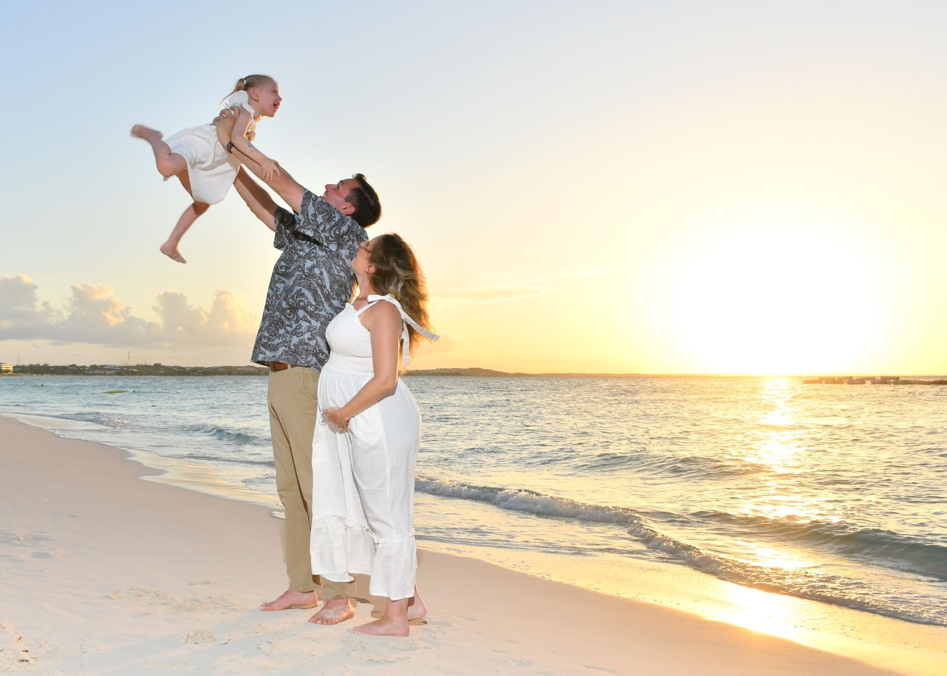 Family playing on a beach at sunset, with a child lifted into the air near the water.