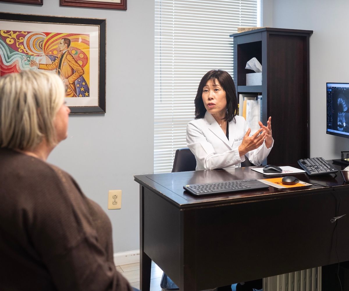 Two people talking across an office desk, with a computer and framed art in the background.