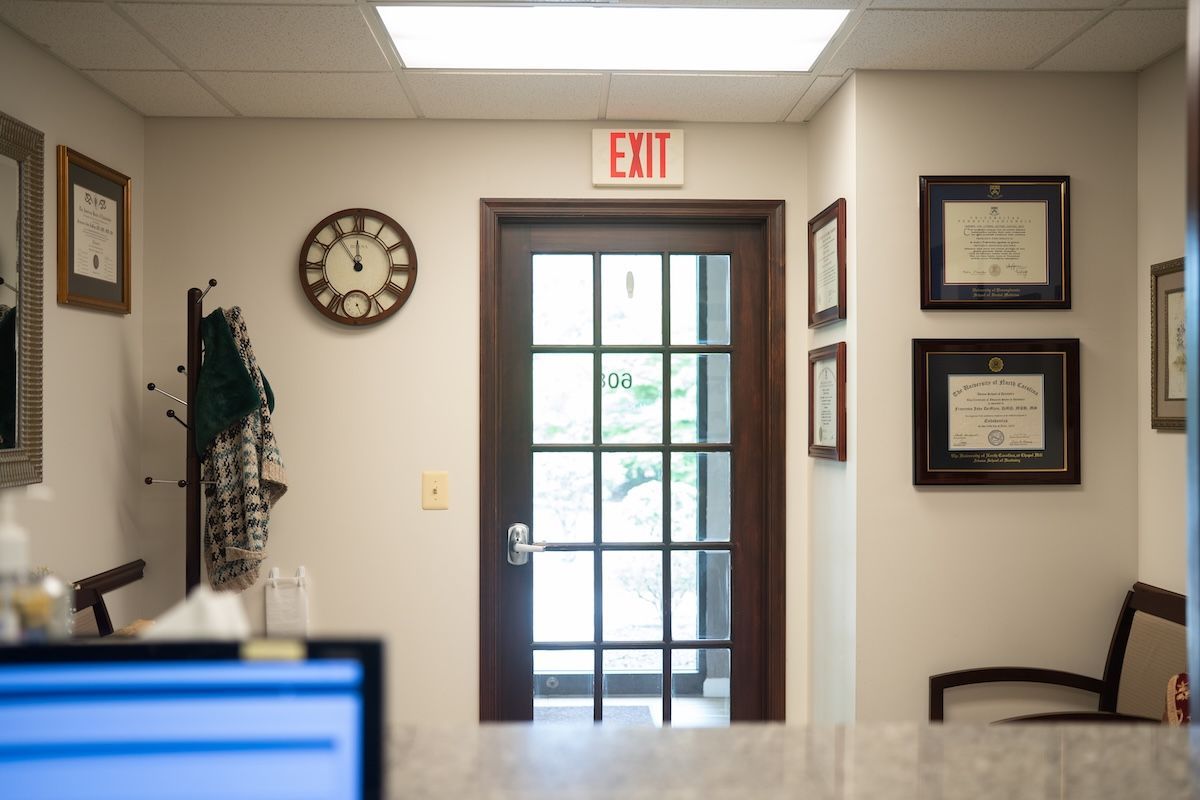 Office hallway with exit door, wall clock, framed certificates, and a chair near the wall.