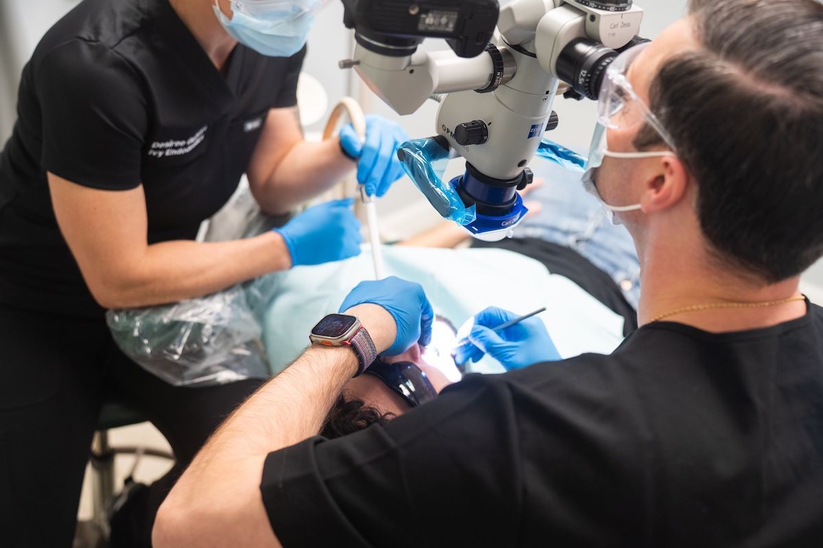 Dental team performing a procedure under a microscope, with gloved hands and a patient reclined on the chair