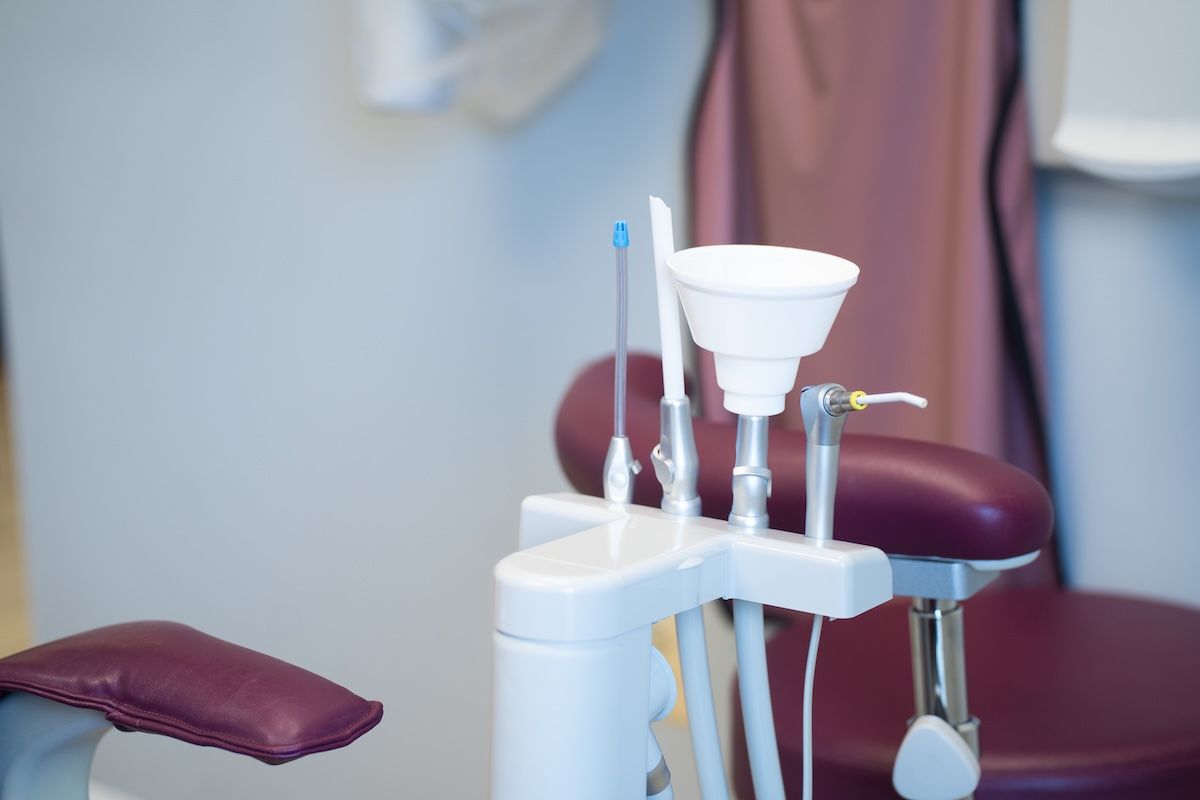 Dental clinic chair with maroon cushions, tray, and tools near a sink.