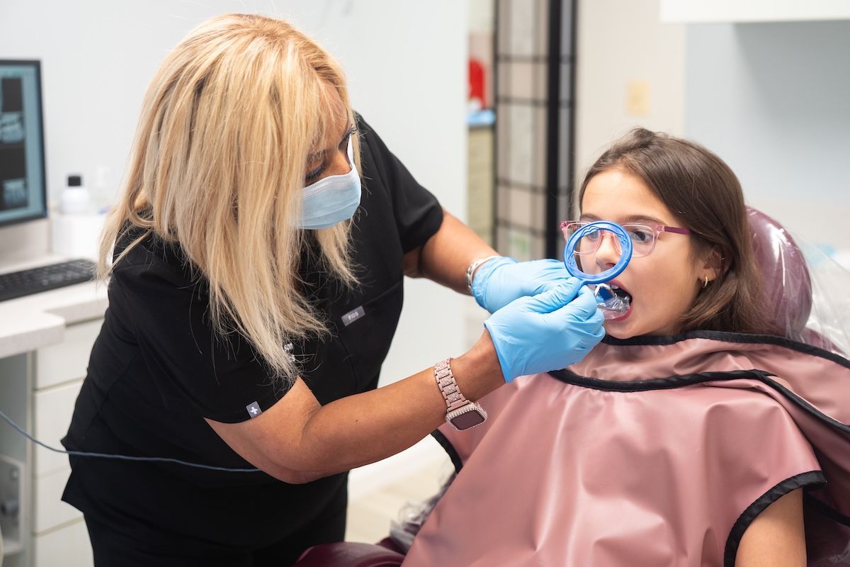 Dentist examining a patient’s teeth with a mirror in a clinic, both wearing face masks and gloves