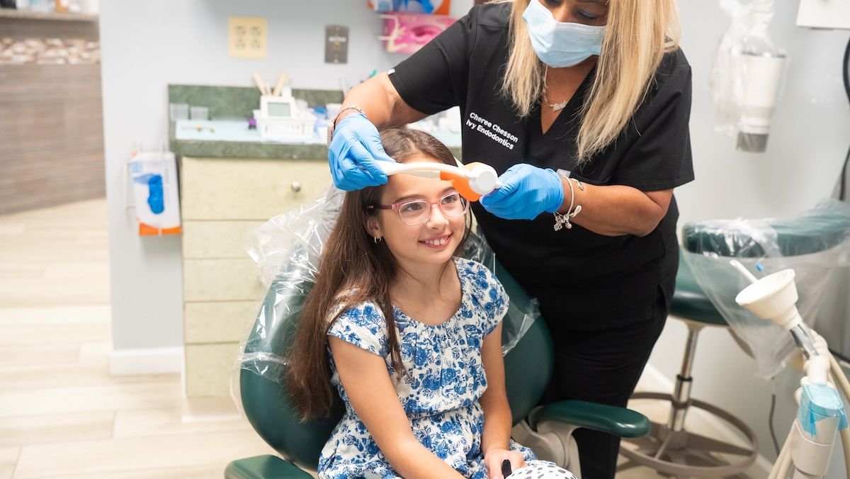 Masked dentist examining a child patient in a dental chair while holding a bright blue tool