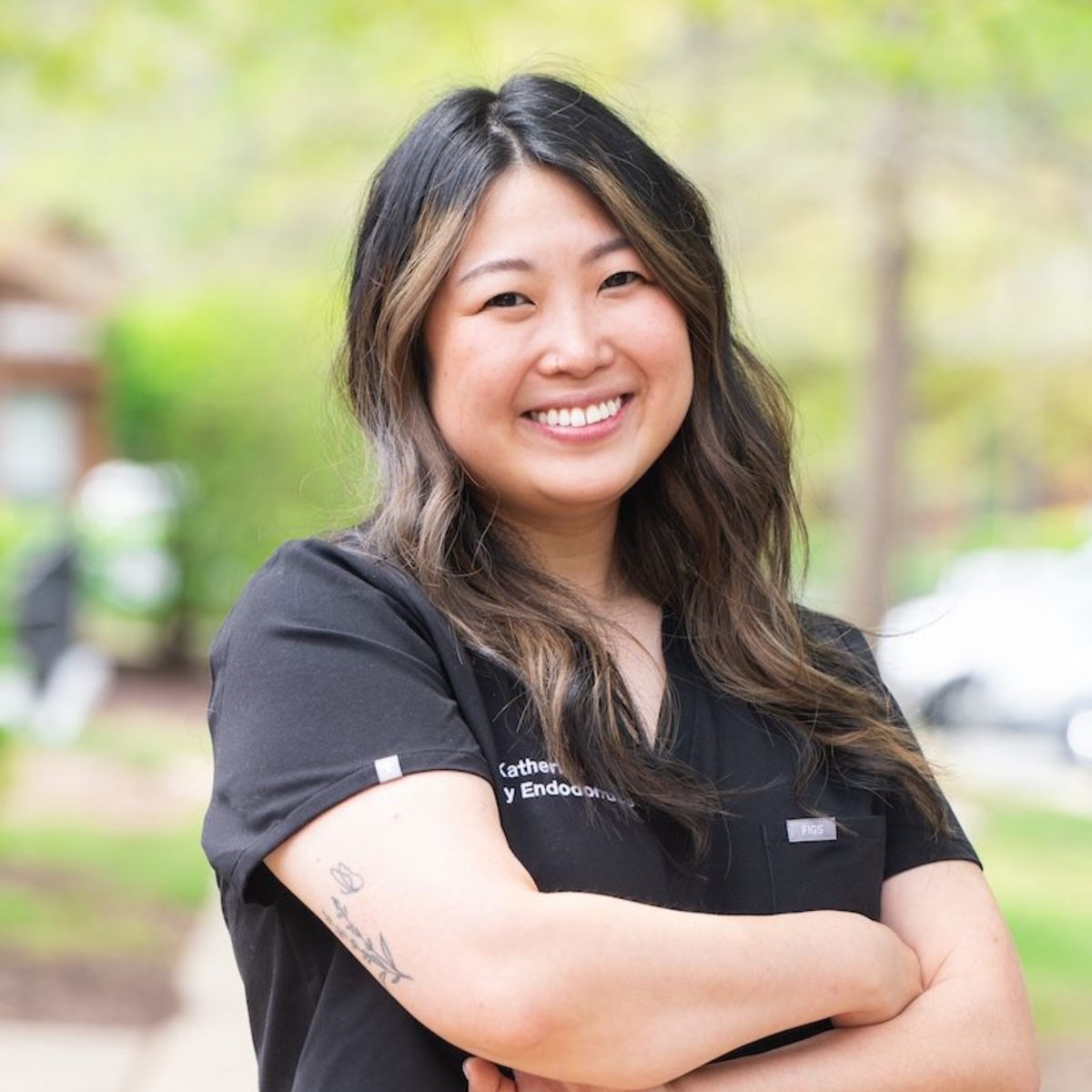 Smiling person with long dark hair, arms crossed, wearing a black shirt outdoors.