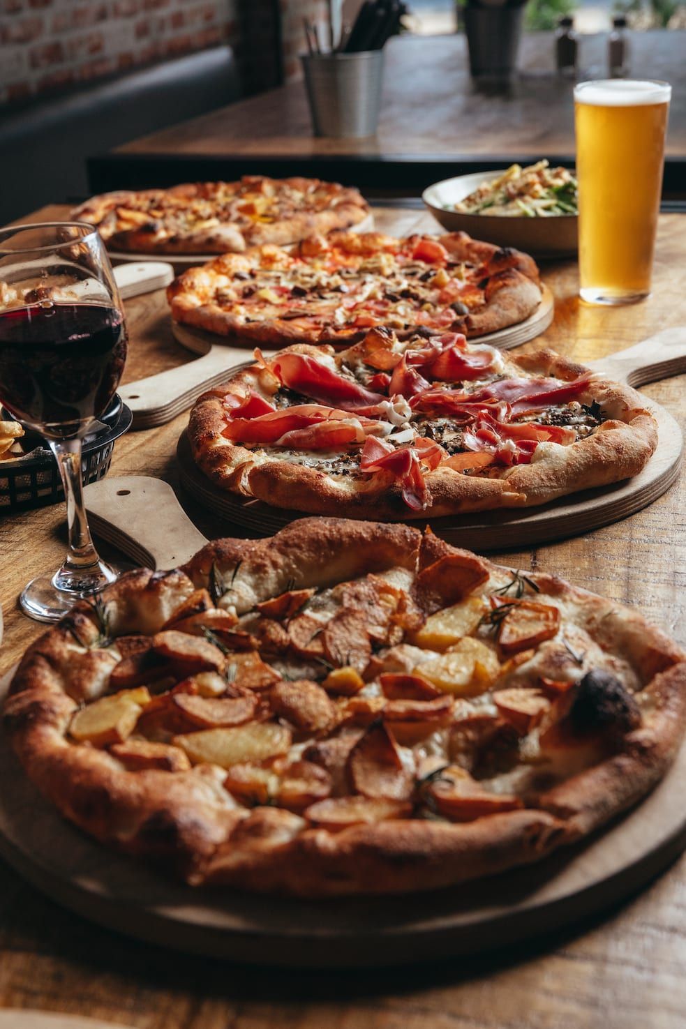 A table topped with three pizzas , a glass of wine and a glass of beer.