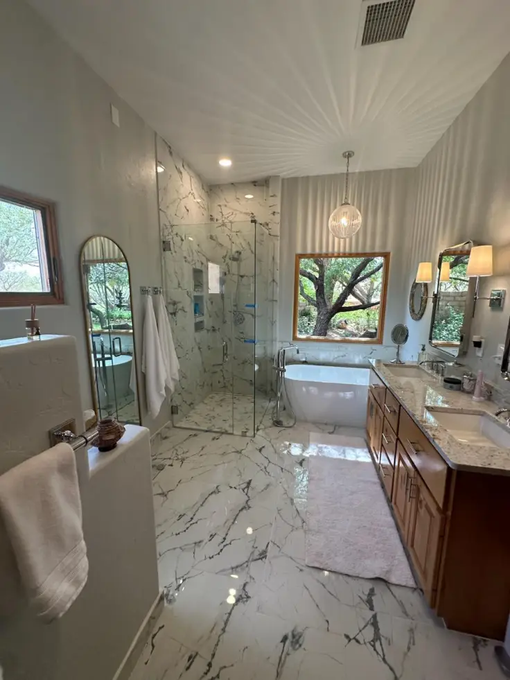 Modern bathroom with marble floors, glass shower, freestanding tub, and vanity under a large window