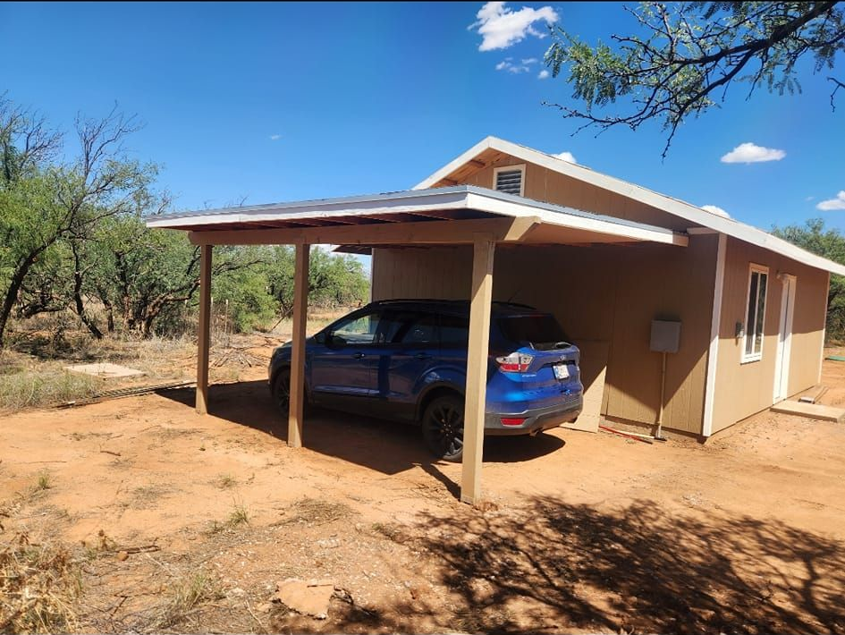 Blue car parked under a small carport beside a tan house in a dry, rural landscape.