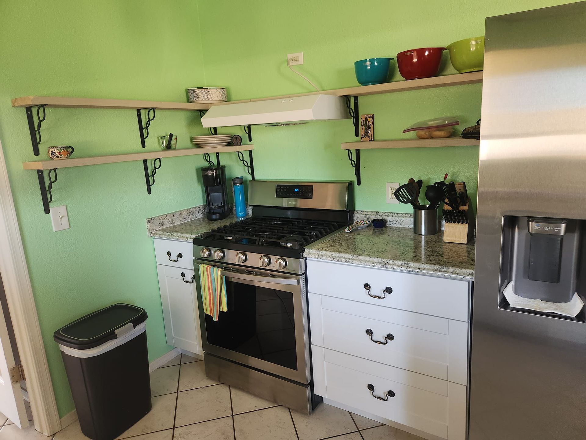 Small kitchen with mint-green walls, open shelves, stove, countertop, sink, and stainless steel refrigerator.