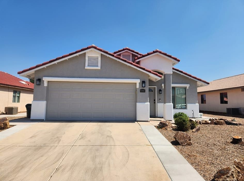 Single-story gray house with a two-car garage, red roof, and desert landscaping under a clear sky