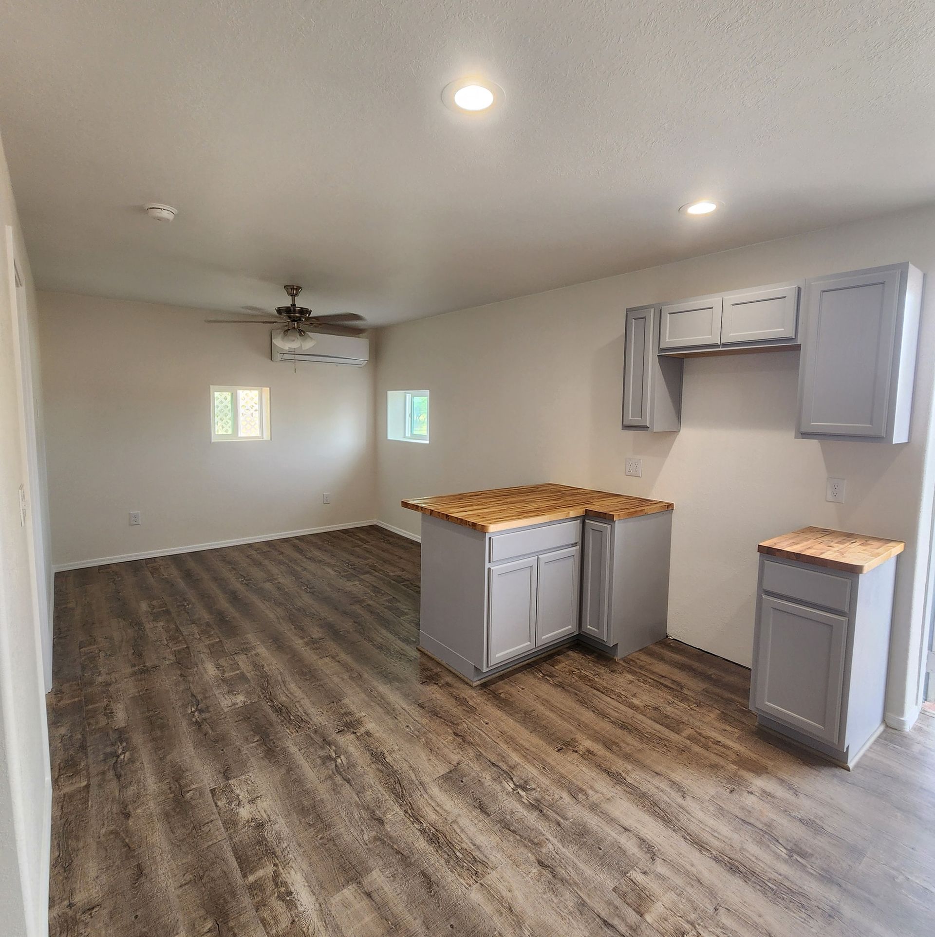 Empty open-plan room with wood floors, white walls, ceiling fan, and a small kitchen island and cabinets.