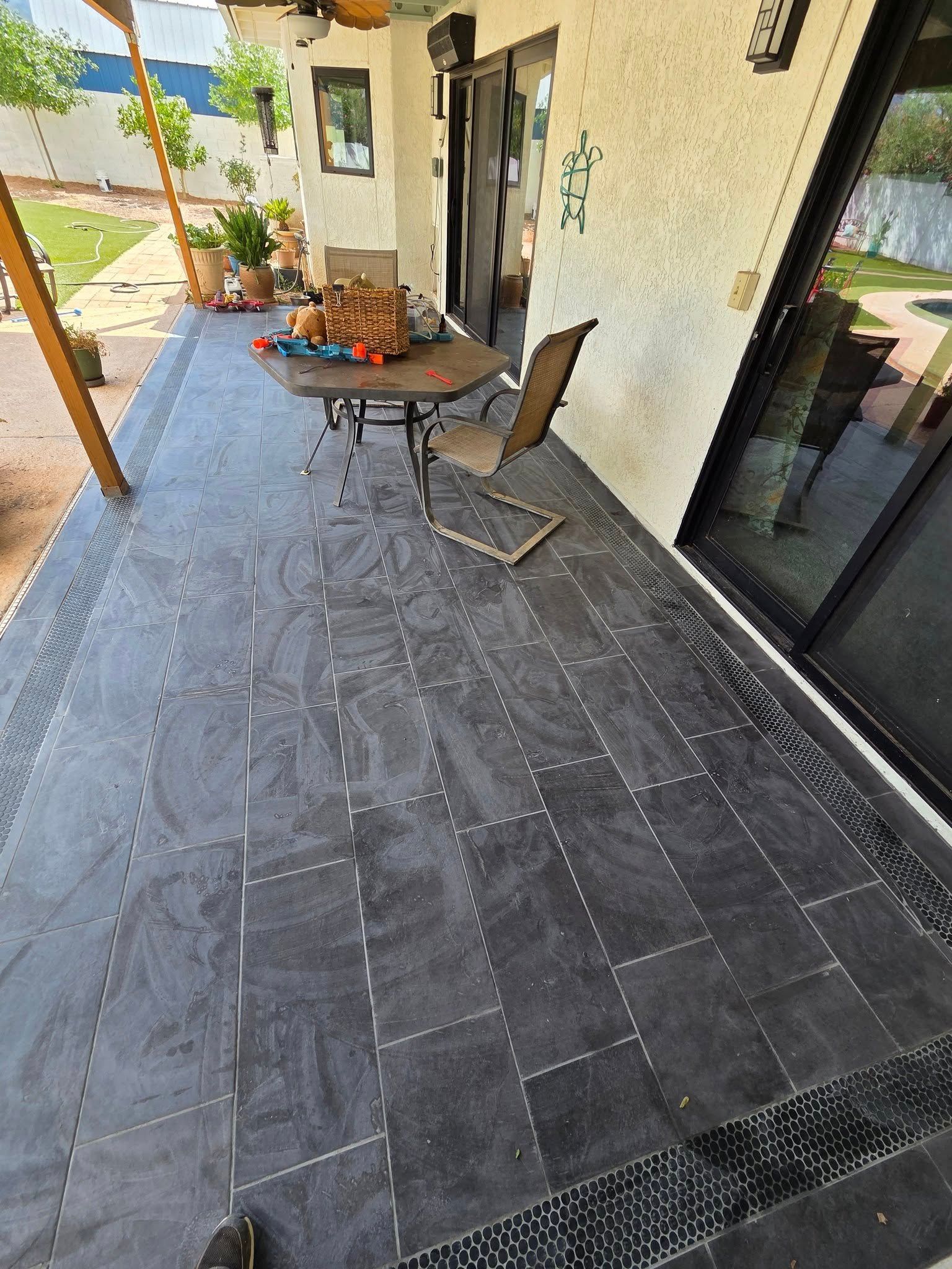 Stone patio walkway beside a house, with a table and chairs and potted plants along the wall.