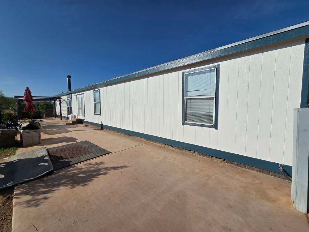Long white mobile home with blue trim beside a concrete patio under a clear blue sky