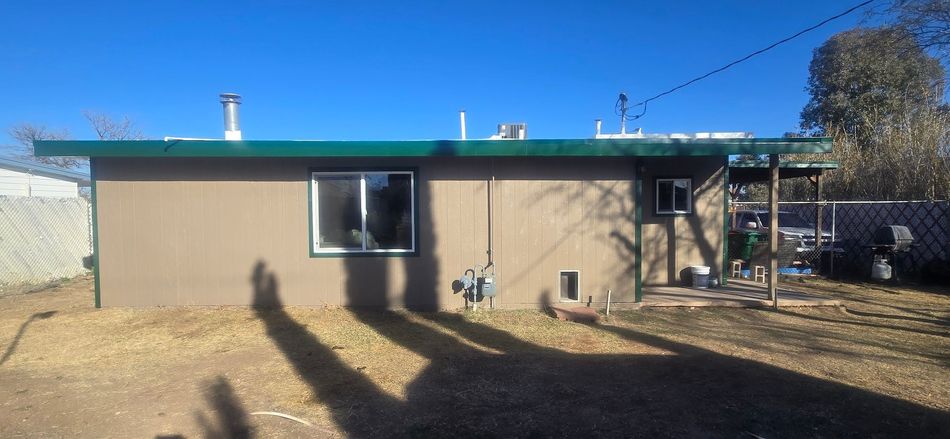 Small tan house with green roof and a window in a sunny yard with long shadows.
