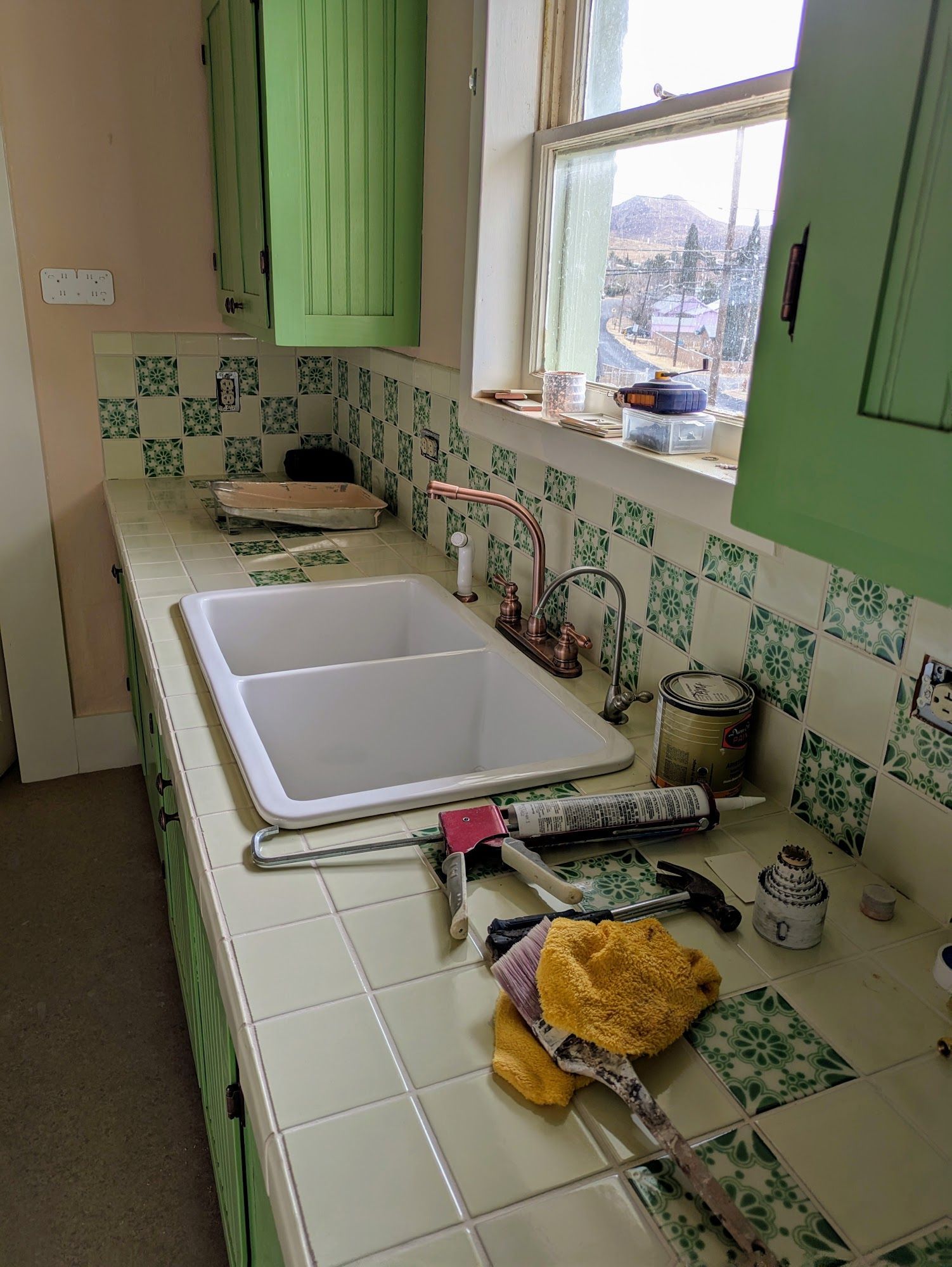 Green kitchen with tiled counter, double sink, and window overlooking a yard.