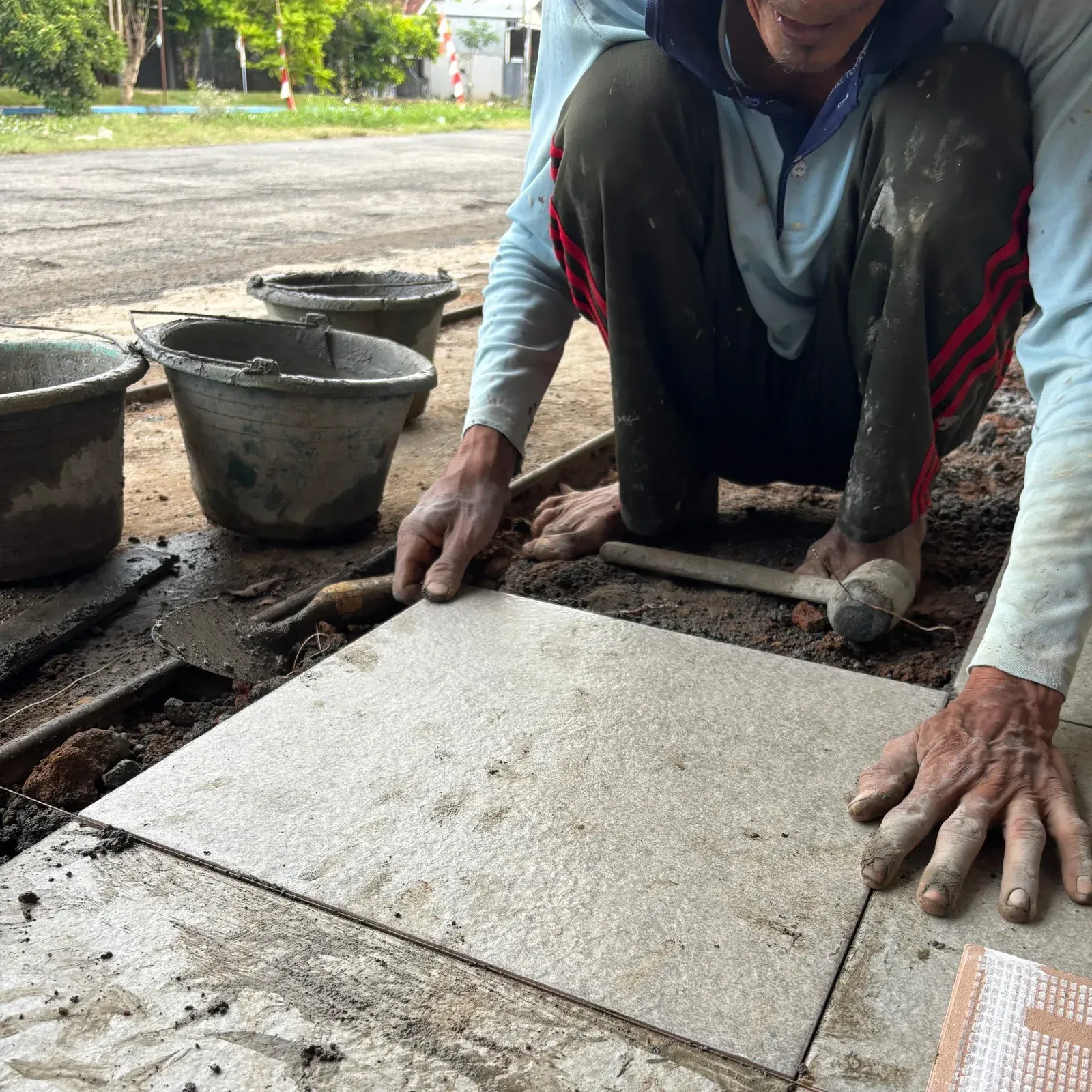 Worker installing concrete tiles on a wet sidewalk, with buckets and tools nearby.