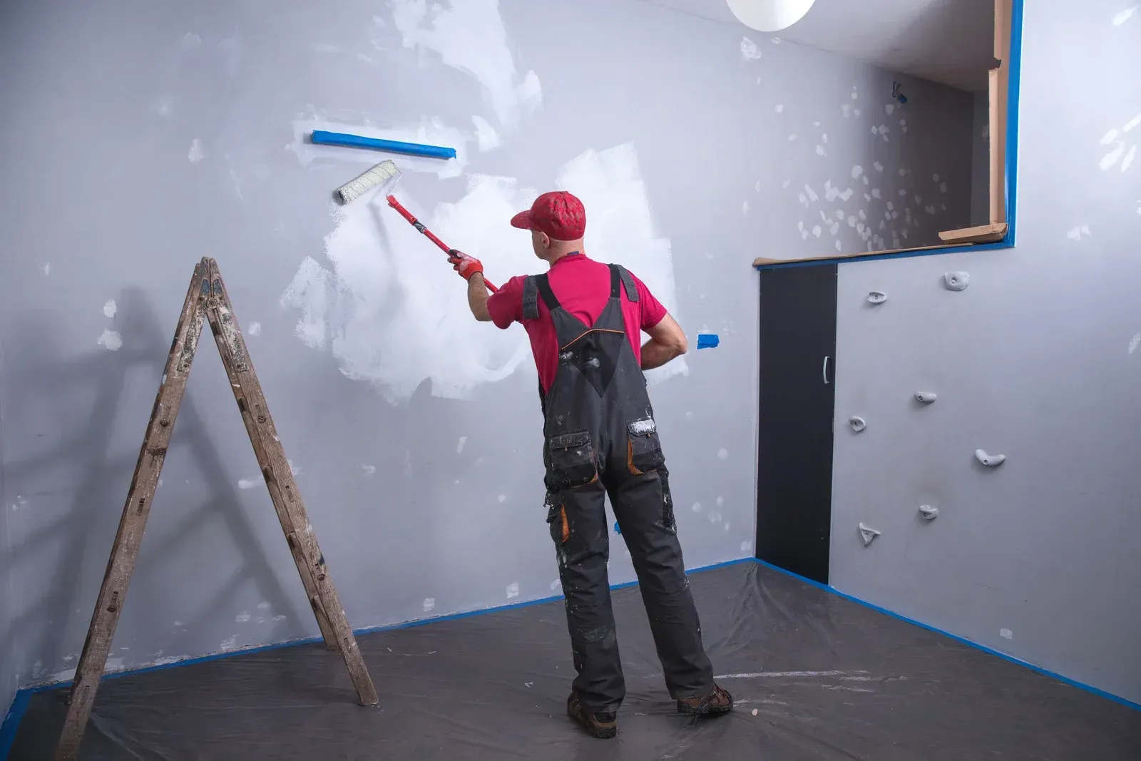 Painter rolling white paint on gray walls in a room with a ladder and blue tape borders