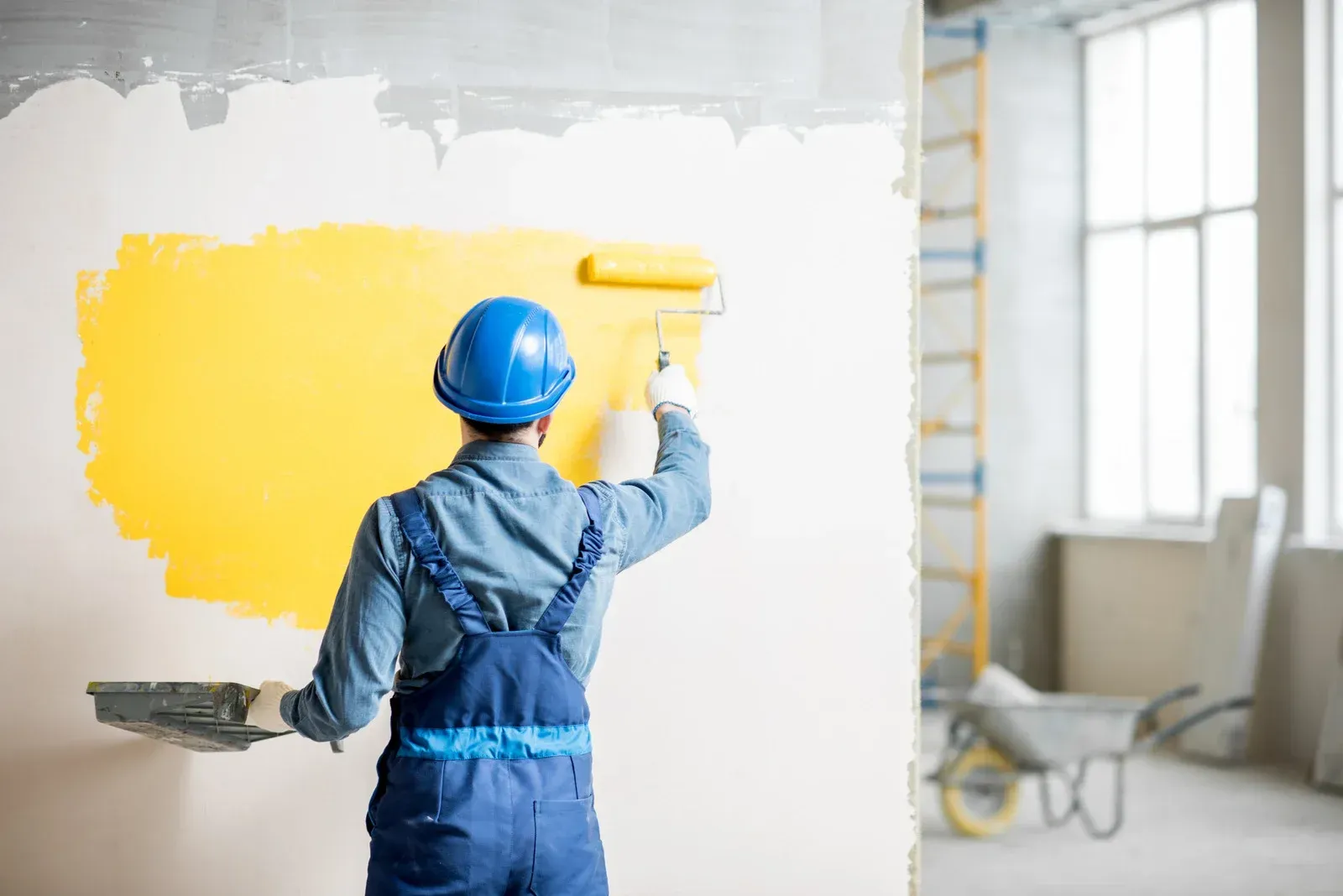 Painter in blue overalls and hard hat applying bright yellow paint to a white wall indoors