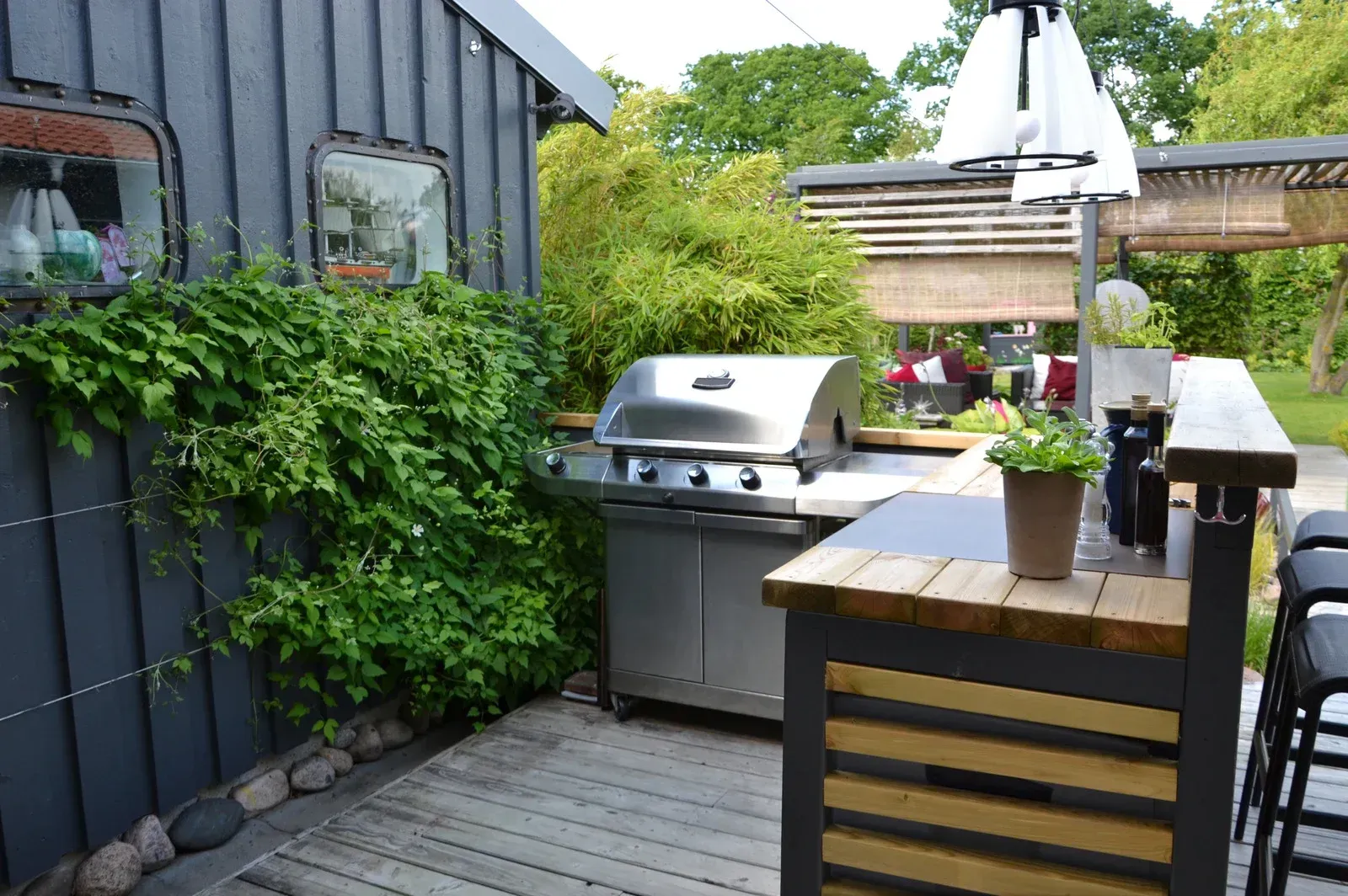 Small backyard patio with a stainless grill, wooden bar table, and potted plants beside a dark shed