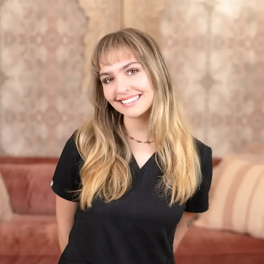 Smiling woman with blonde hair in a black top, standing before a patterned wall and pink sofa
