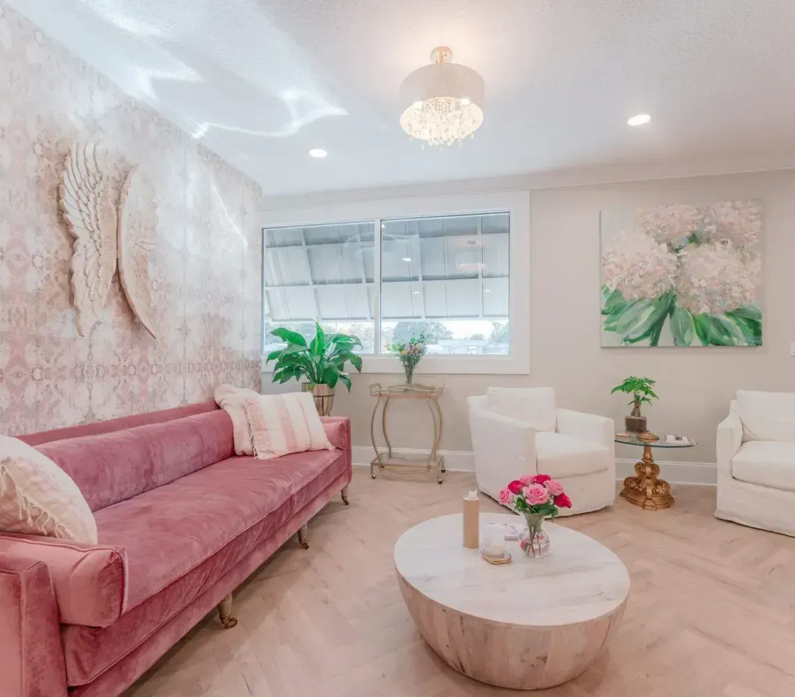 Bright pastel living room with pink sofa, white chairs, coffee table, and floral wall art