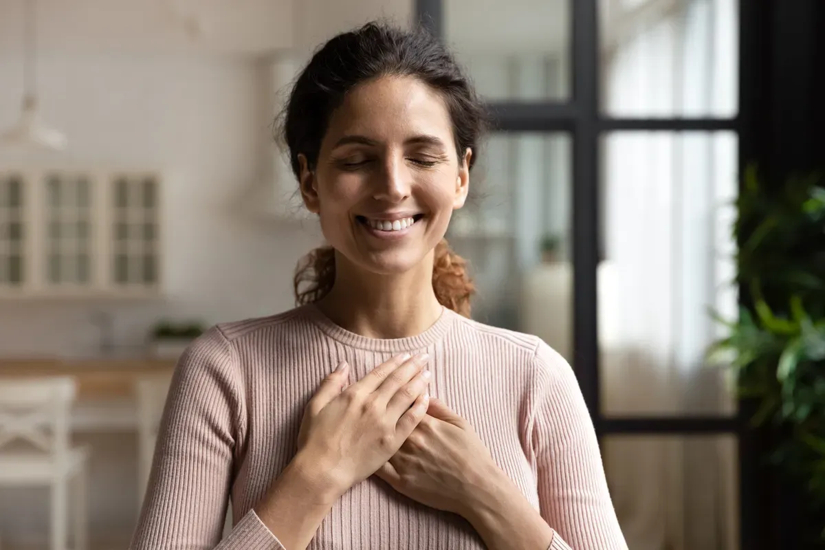 Uma mulher está sorrindo com os olhos fechados e as mãos no peito.
