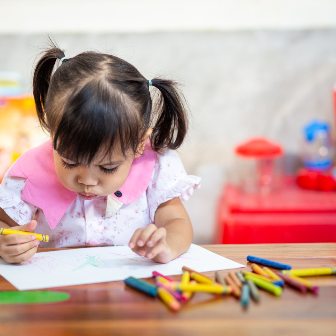 A little girl is sitting at a table drawing with crayons.