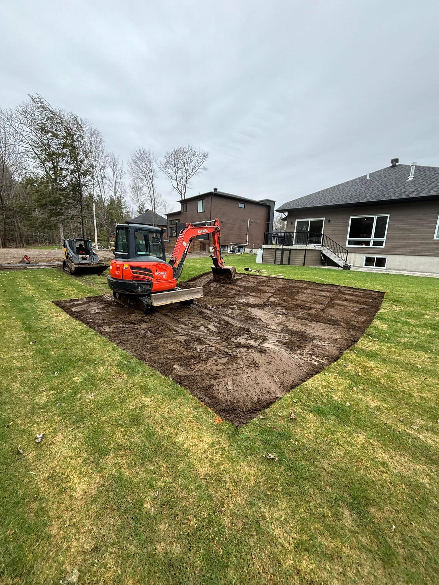 Une petite pelleteuse creuse dans une cour, préparant une zone rectangulaire. Des maisons et des arbres se dressent à l'arrière-plan.
