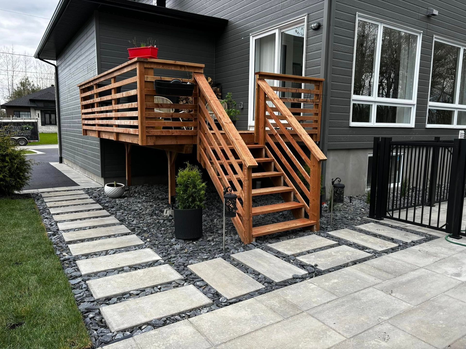 Une terrasse en bois avec des escaliers mène à une maison grise, un chemin de pierre est entouré de gravier noir et d'herbe.