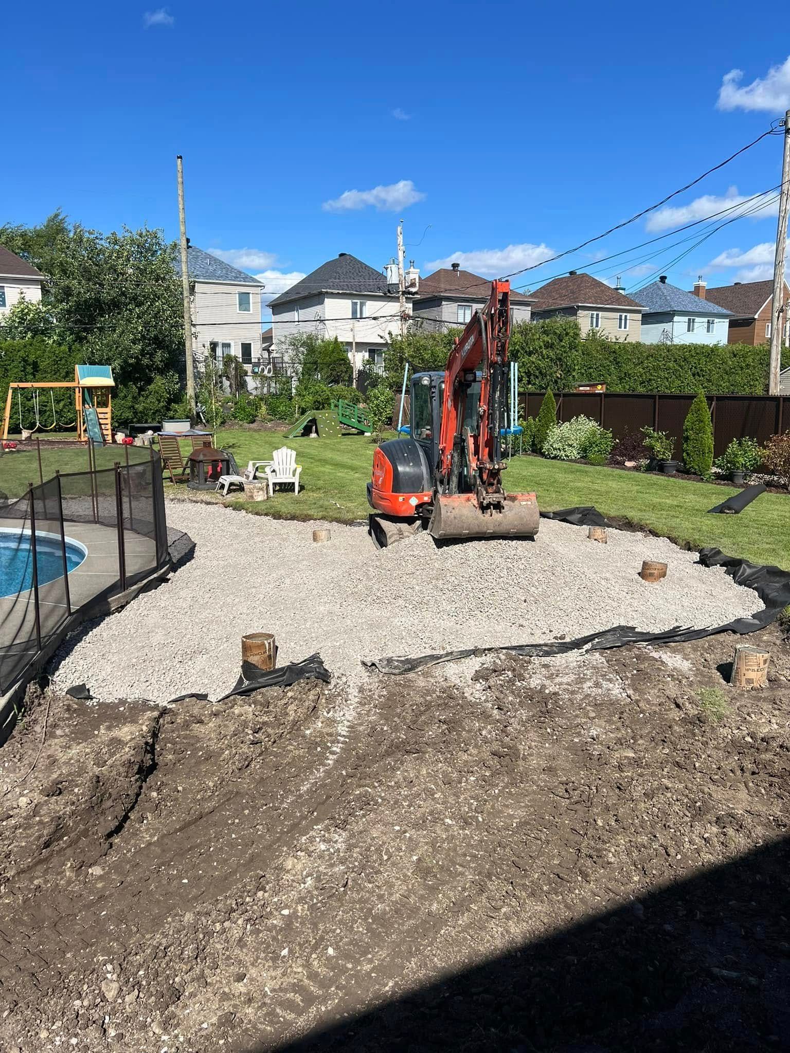 Une petite pelleteuse sur du gravier dans un jardin, près d'une piscine, avec des maisons en arrière-plan.