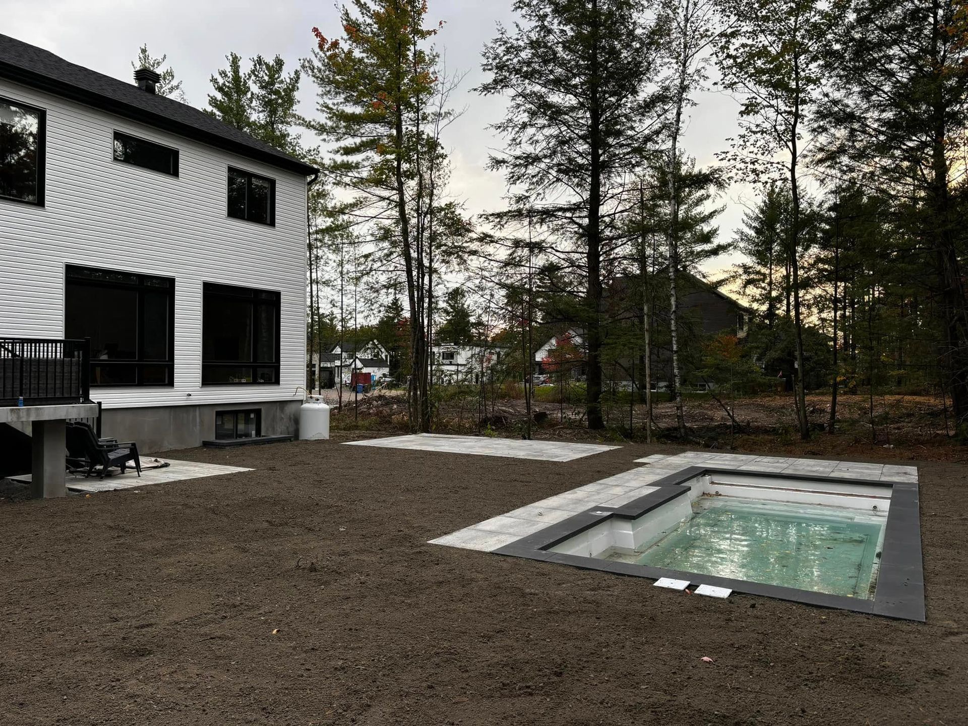 Jardin avec une maison blanche moderne, une petite piscine rectangulaire et un terrain nu. Arbres en arrière-plan.