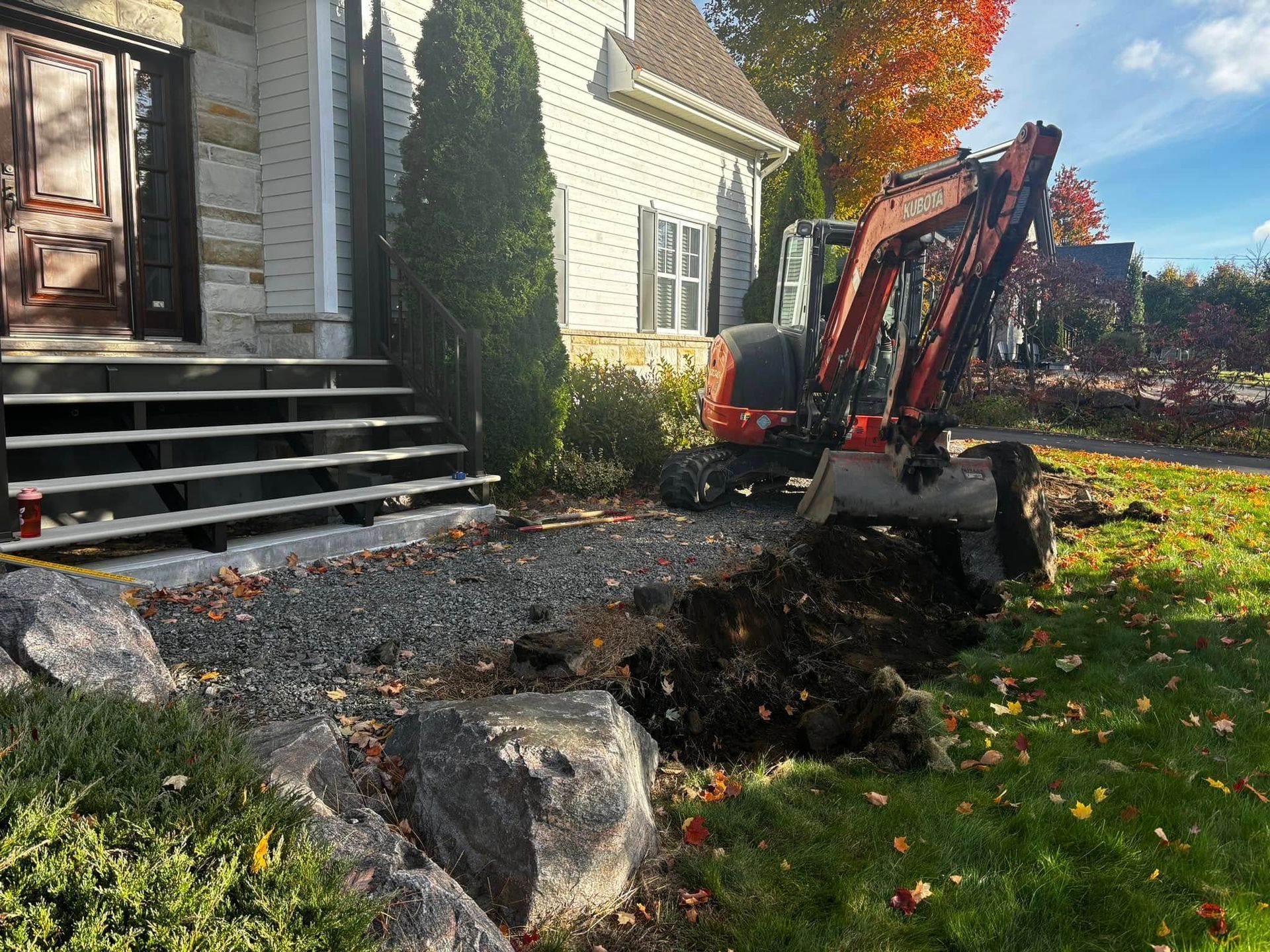 Une excavatrice creuse près du perron d'une maison, dans un sol sombre, parsemé de pierres et de feuillage d'automne.