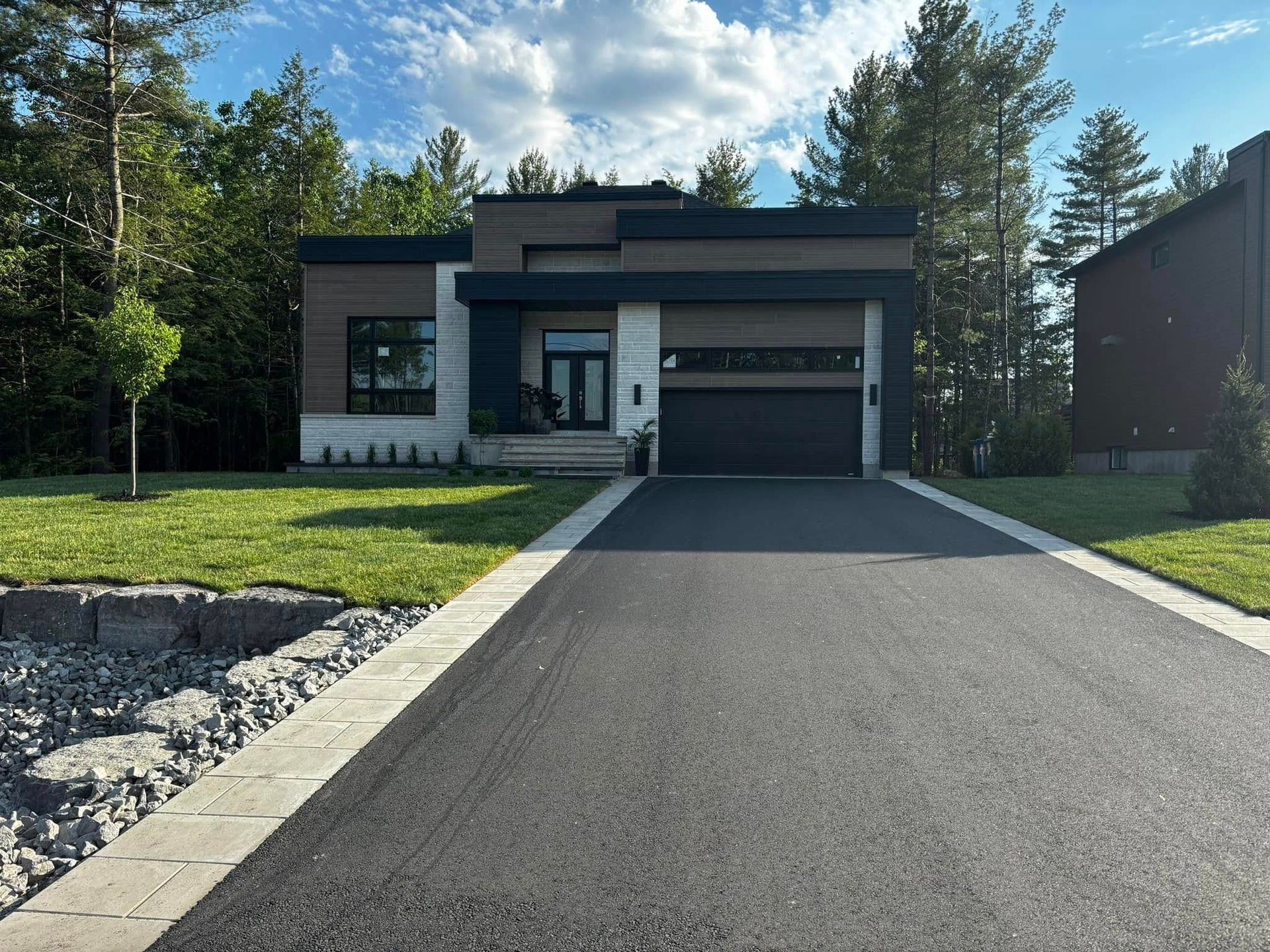 Maison moderne avec porte de garage gris foncé, façade en pierre claire et longue allée en asphalte noir.