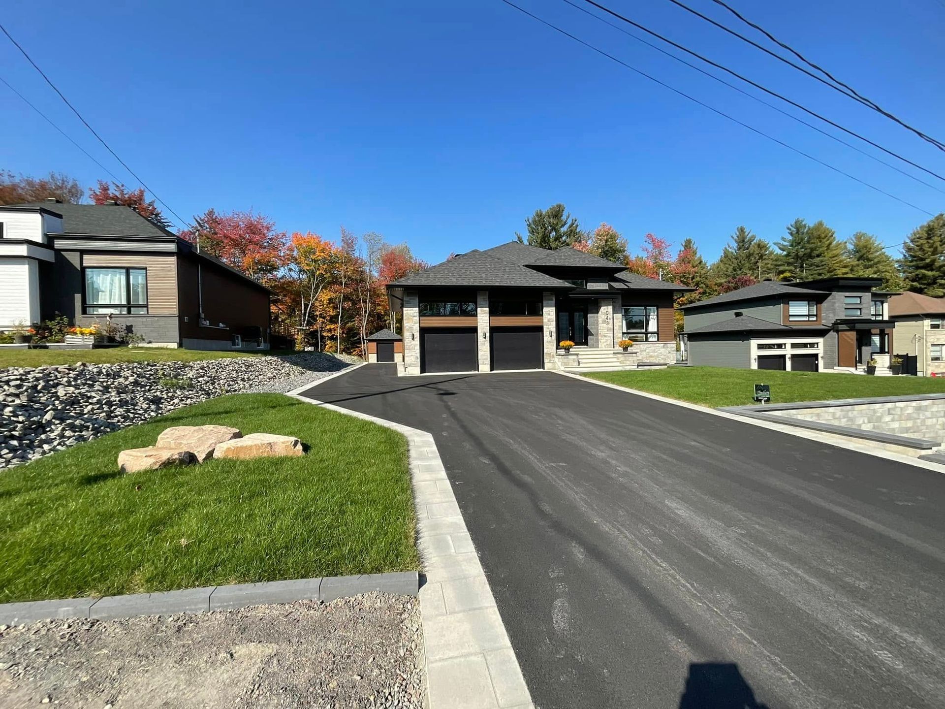 Allée asphaltée menant à une maison moderne avec porte de garage noire, revêtement en pierre et gris, et pelouse bien entretenue.