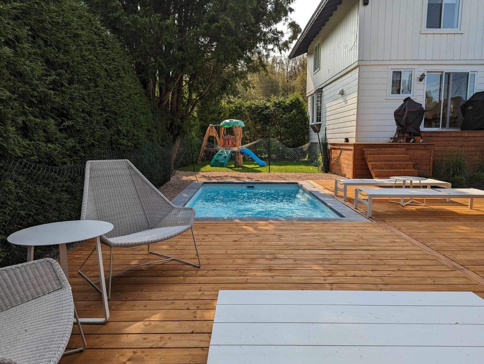 Terrasse en bois avec petite piscine, chaises et structure de jeux dans un jardin près d'une maison blanche.