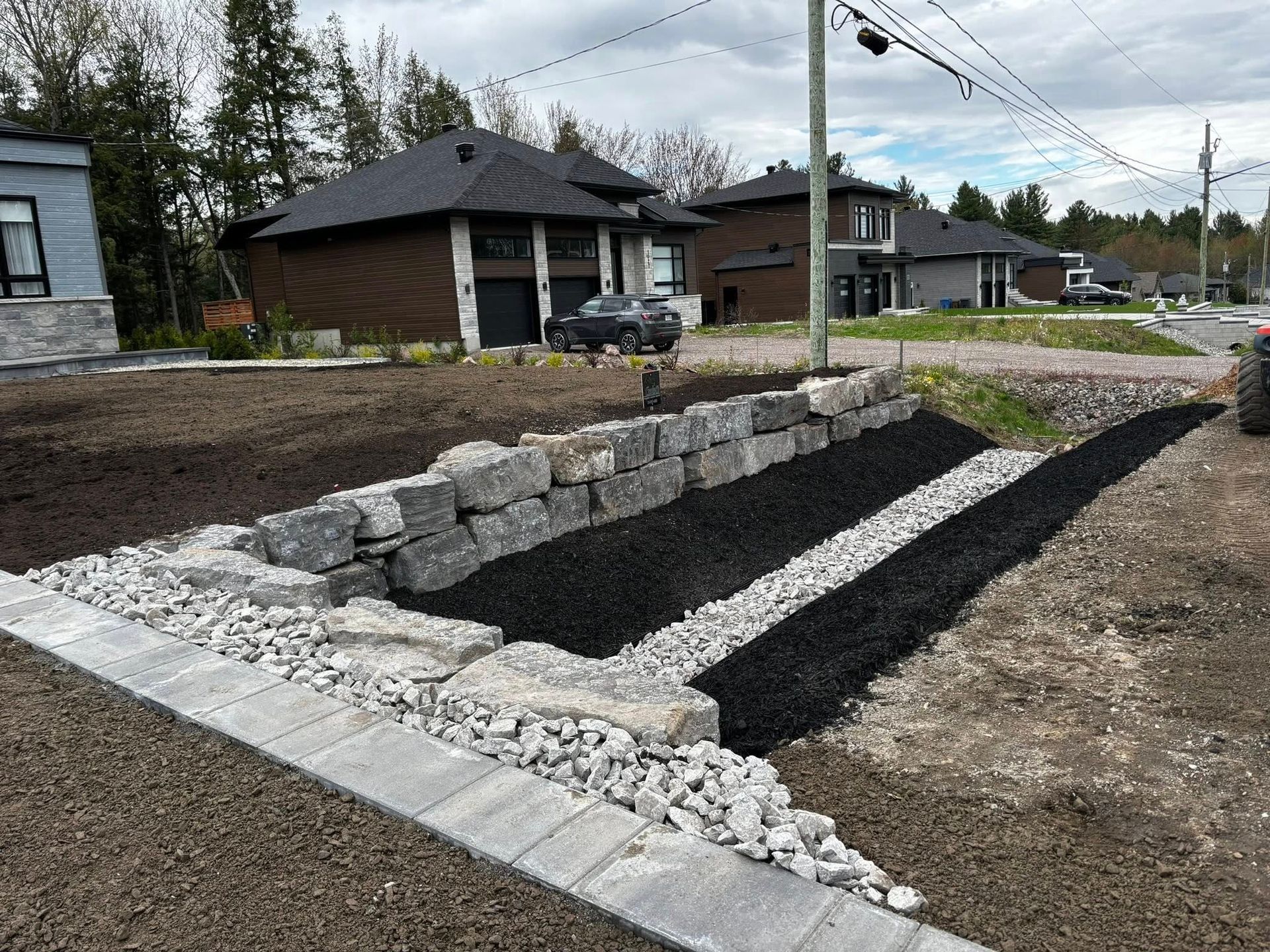 A constructed landscape with retaining walls and gravel. Dark mulch and grey stone are visible, with houses in the background.