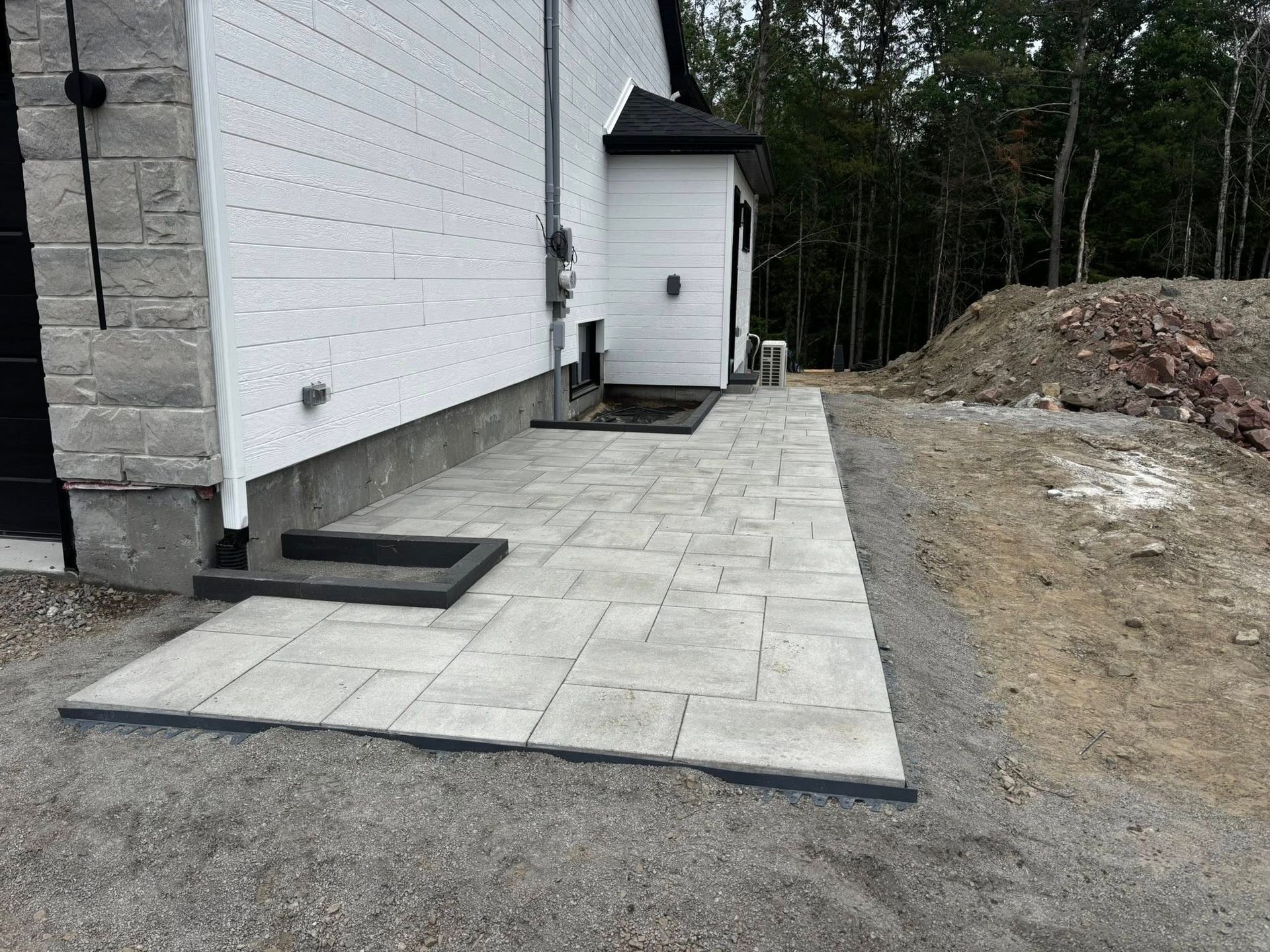 Terrasse en pierre jouxtant une maison blanche aux boiseries sombres. Chemin de gravier et forêt en arrière-plan.