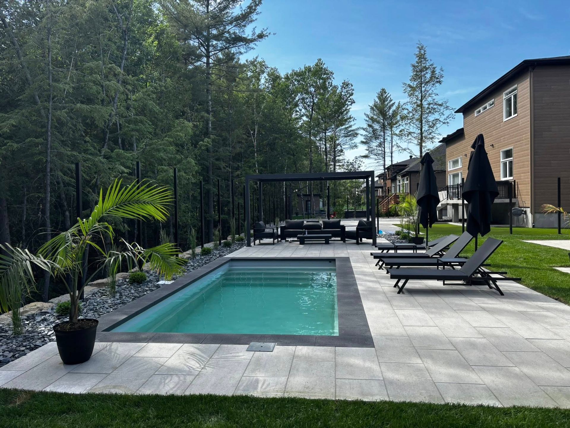 Une piscine rectangulaire entourée de dalles de terrasse, de chaises longues et d'un coin salon avec une pergola dans un jardin.