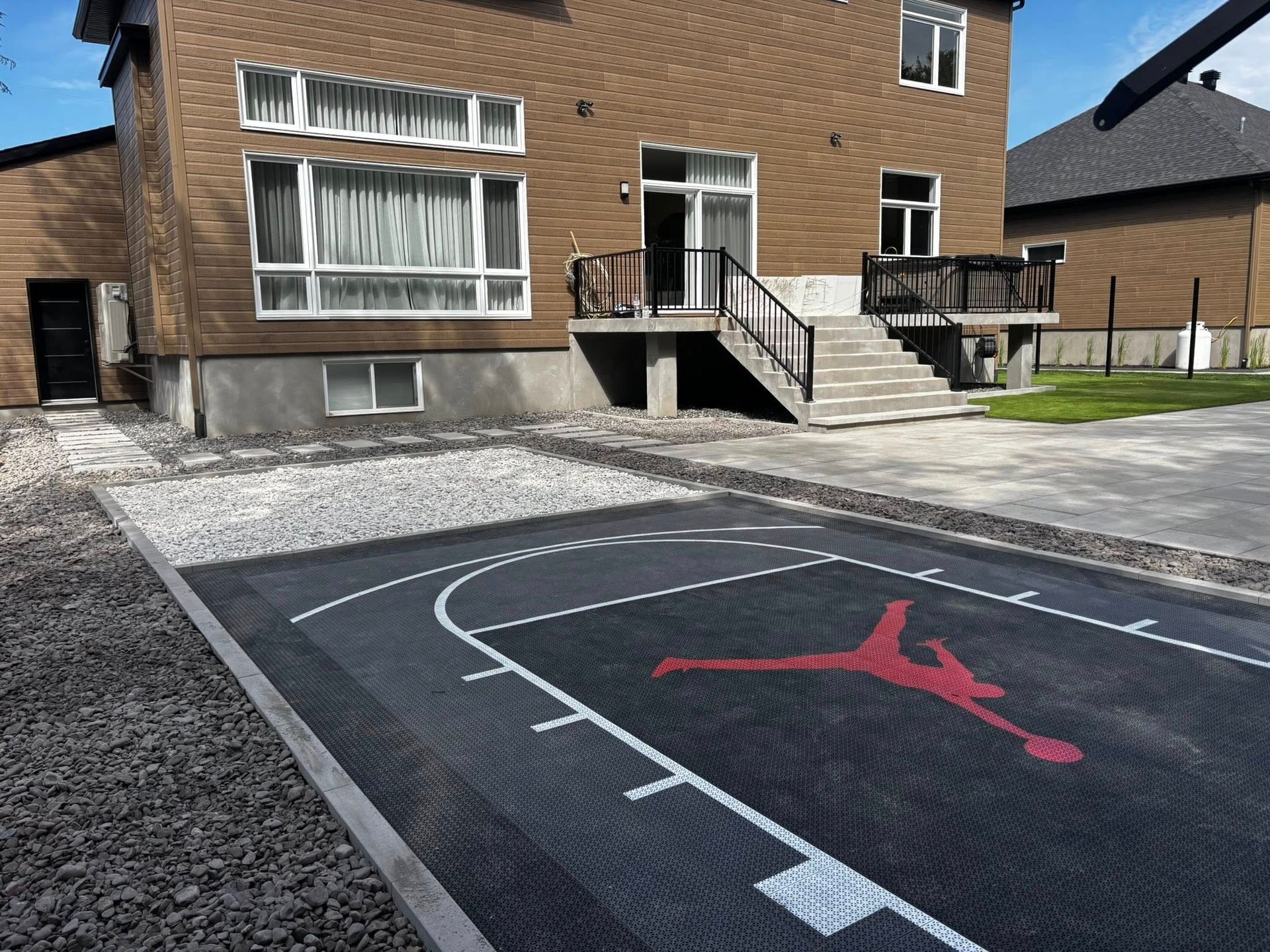 Terrain de basket extérieur orné du logo Air Jordan, à côté d'une maison à l'extérieur marron et bordée de gravier.