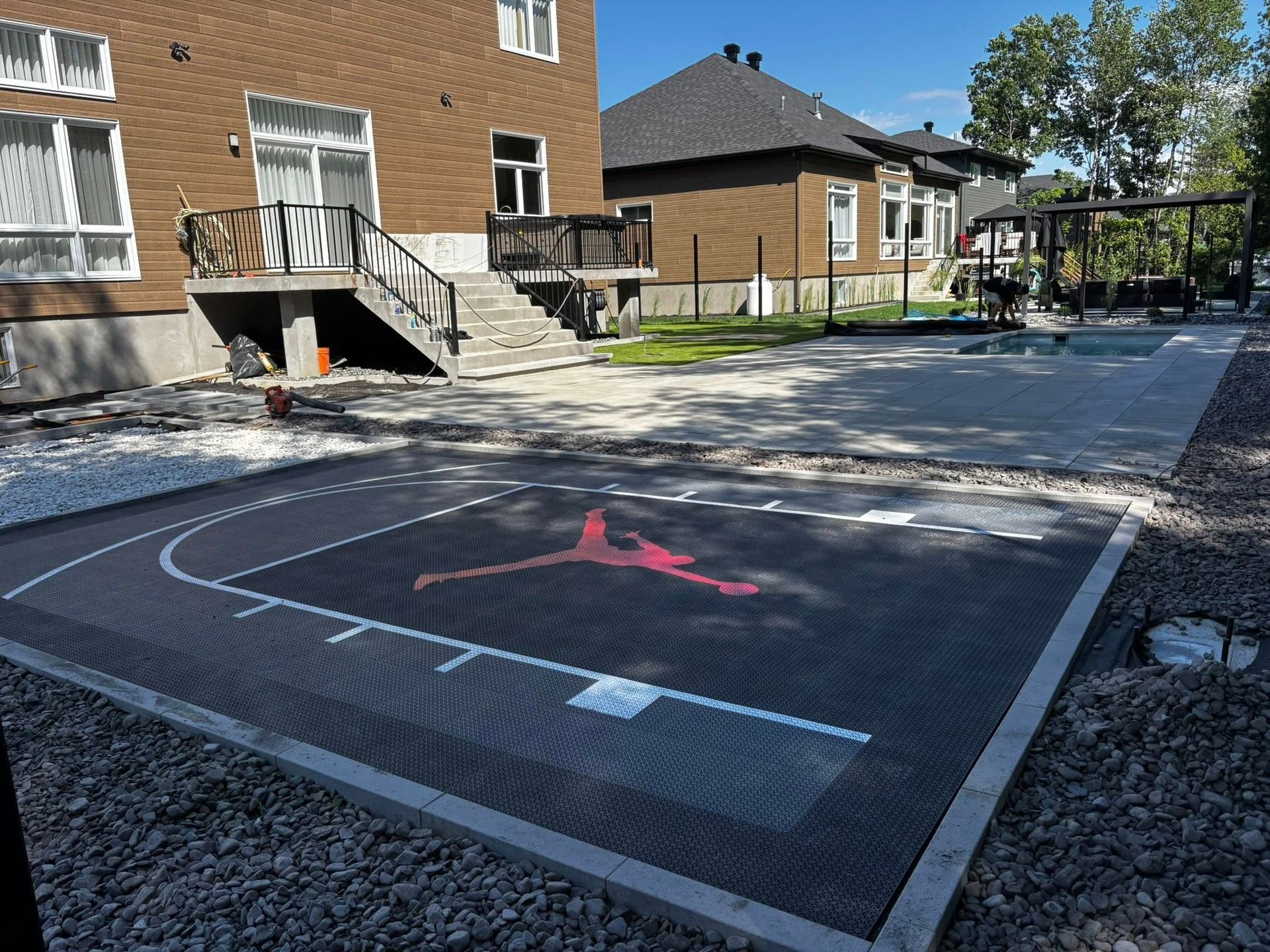 Terrain de basket avec le logo de Michael Jordan dans le jardin. Surface noire, bordure en pierre, maison en arrière-plan.