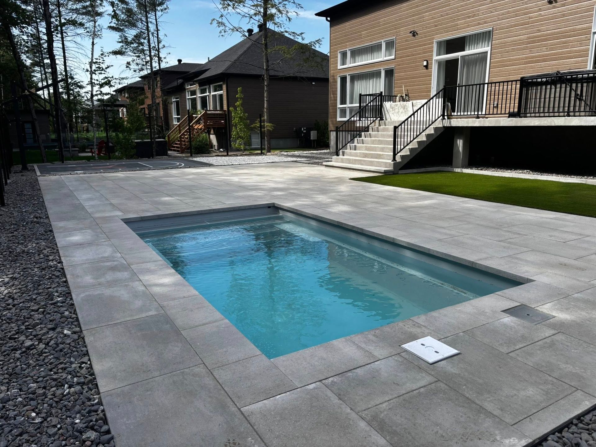 Piscine rectangulaire dans un jardin entouré de pierres grises. Une maison en briques se dresse à l'arrière-plan.
