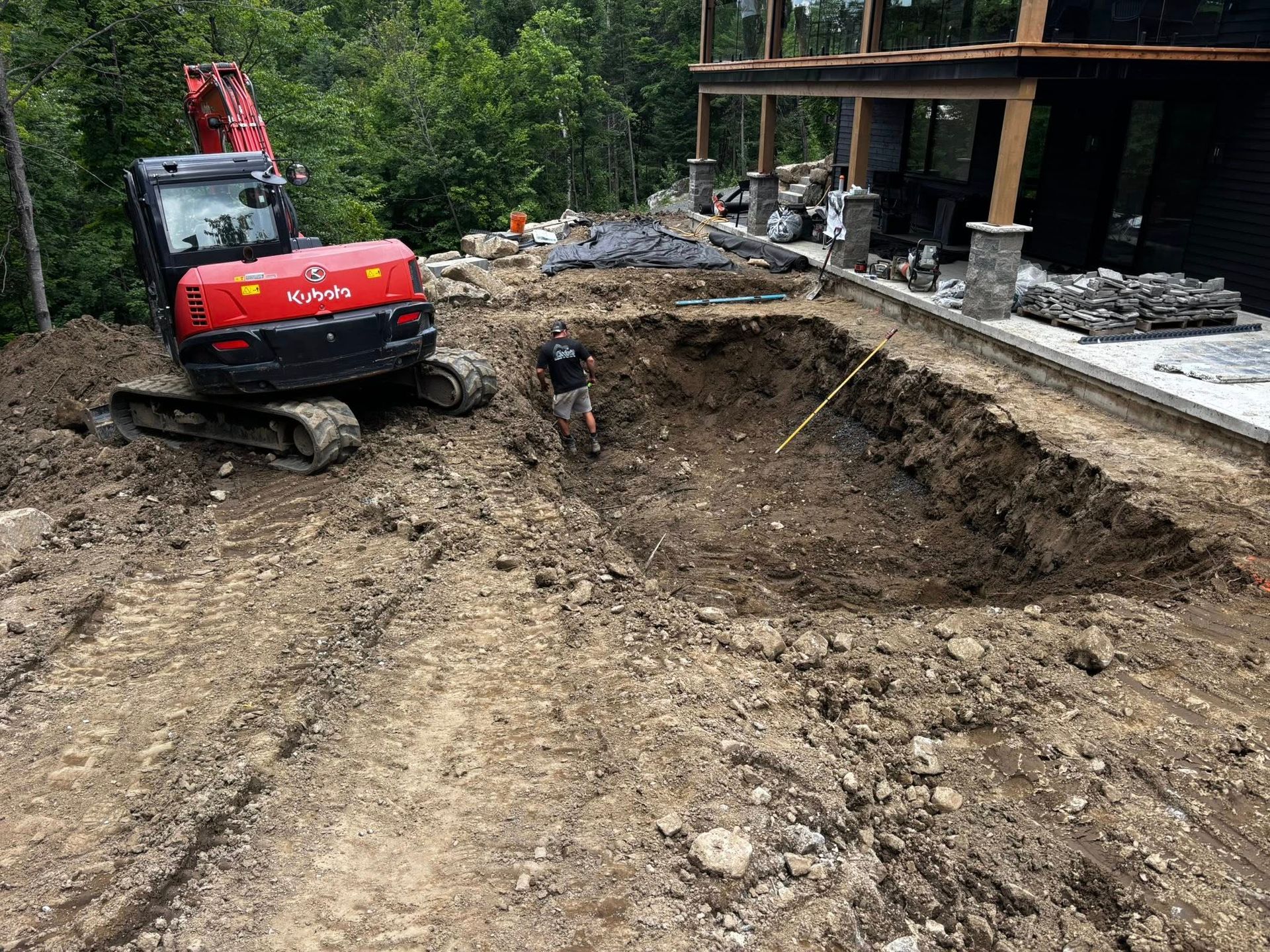Une excavatrice creuse un trou rectangulaire dans un sol boueux près d'un bâtiment.