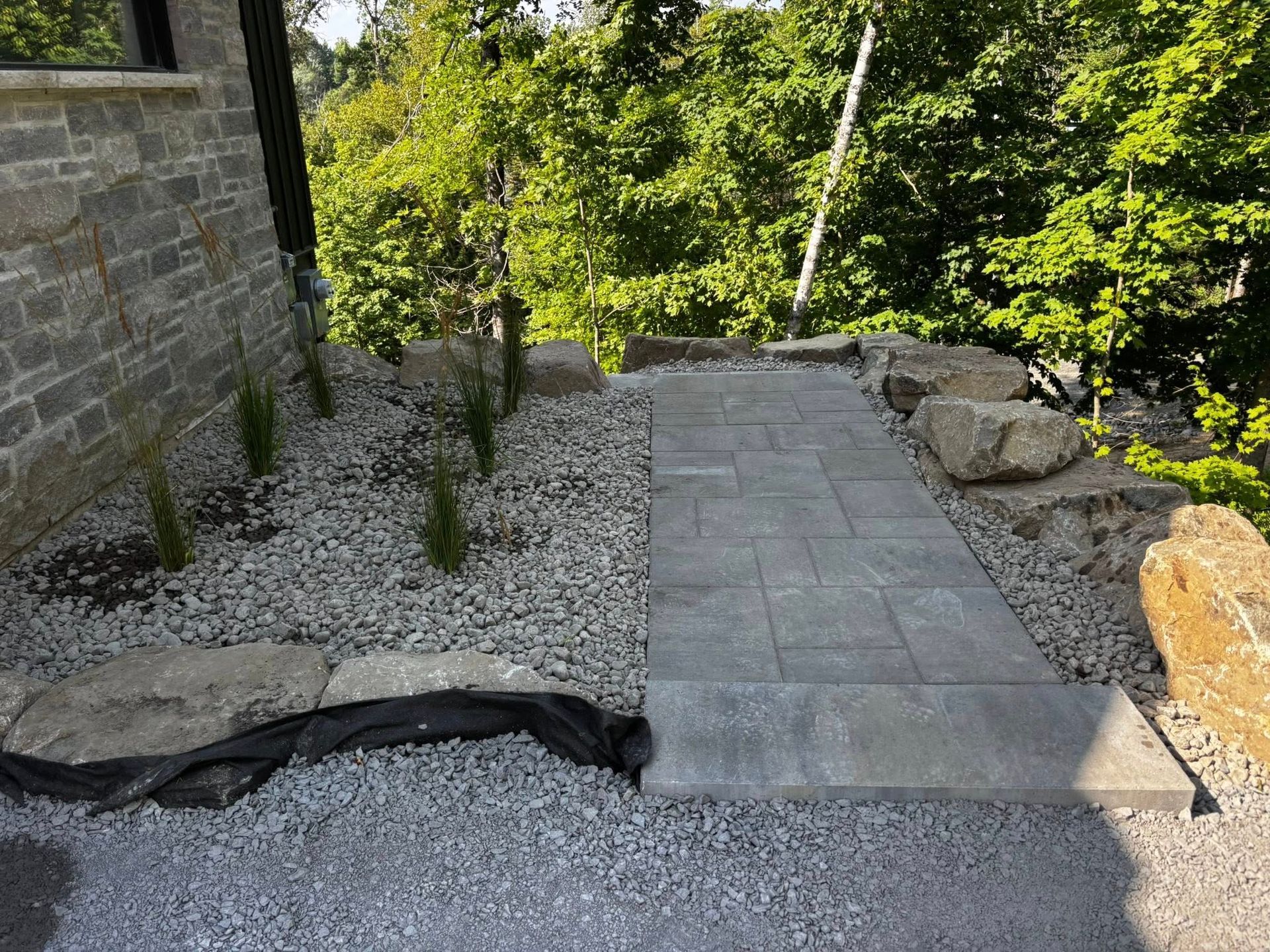 Allée en pierre longeant un chemin de gravier et un bâtiment, avec de gros rochers et de la verdure.