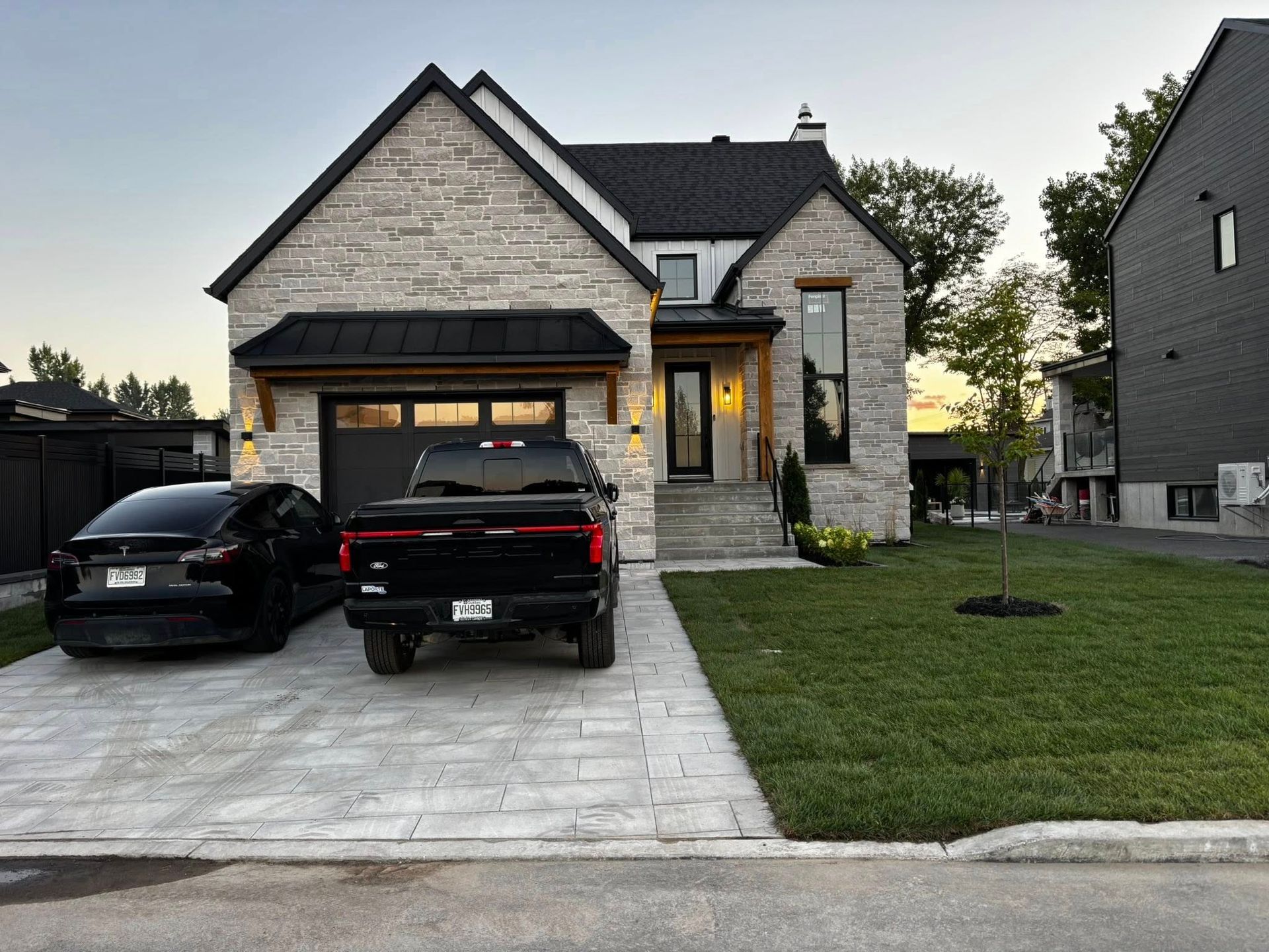 Maison à façade en pierre, garage noir et deux véhicules noirs garés devant, sur une allée pavée.