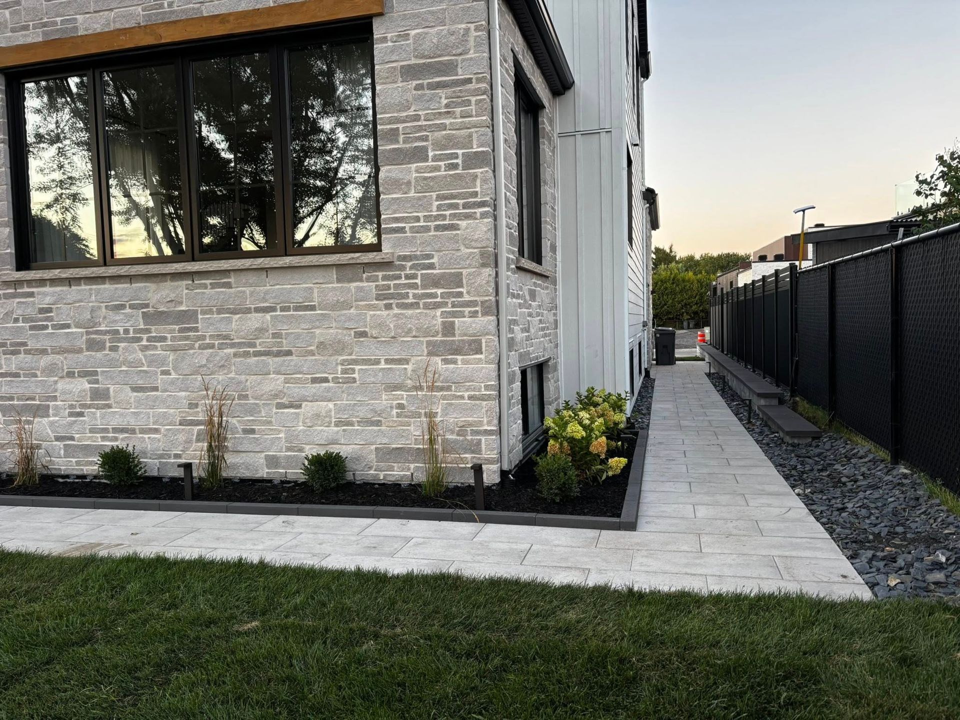 Coin d'une maison moderne avec façade en pierre, grande fenêtre, allée et clôture noire.
