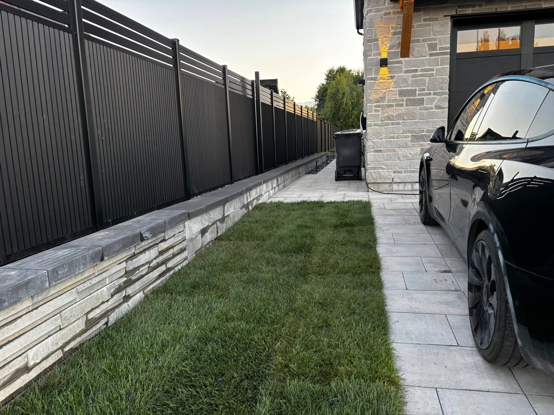 Voiture noire garée à côté d'une allée pavée et d'une zone herbeuse, avec une clôture noire sur le côté.