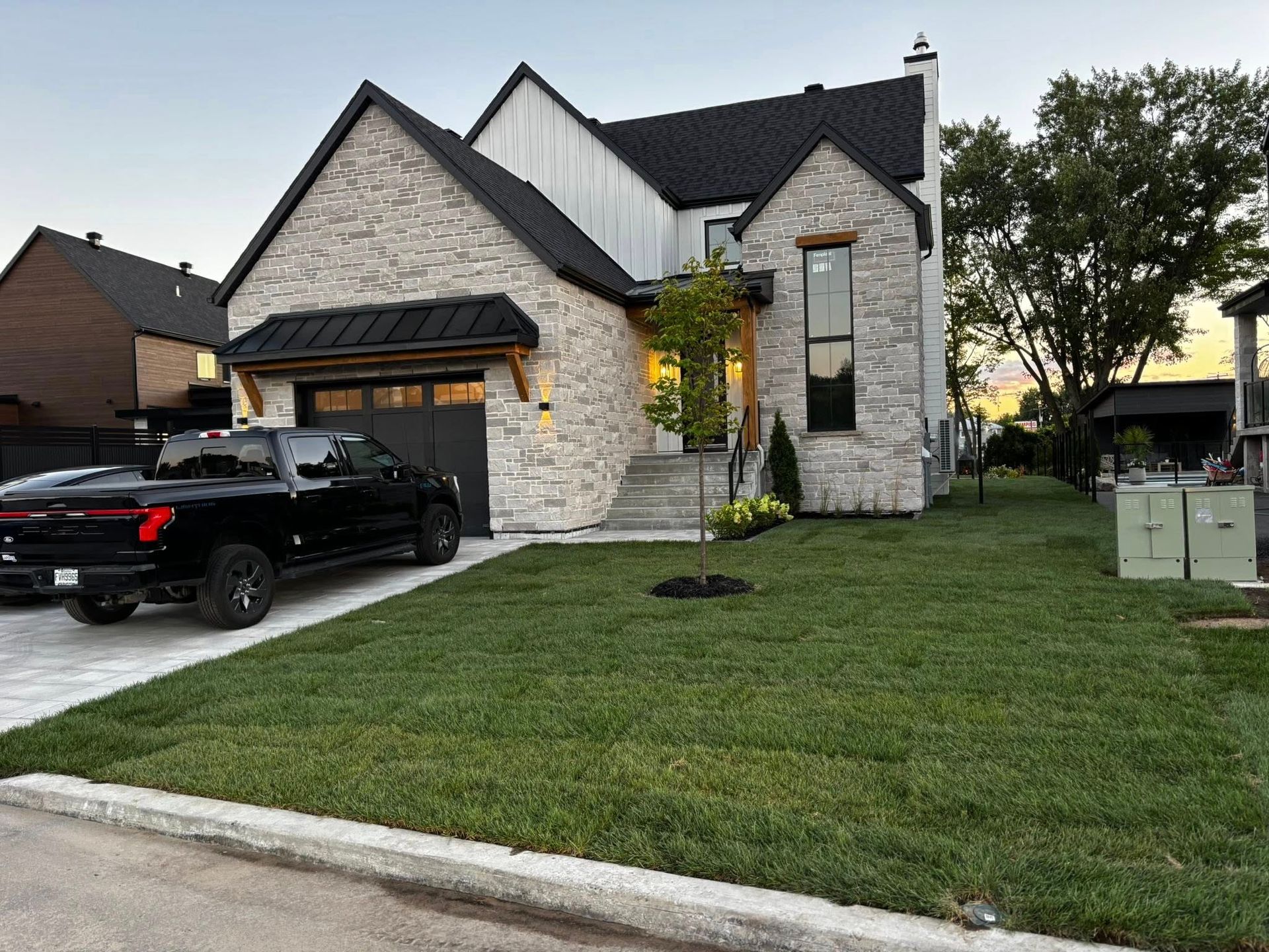 Maison moderne avec façade en pierre, toit noir et pelouse soignée ; camion noir garé dans l'allée.
