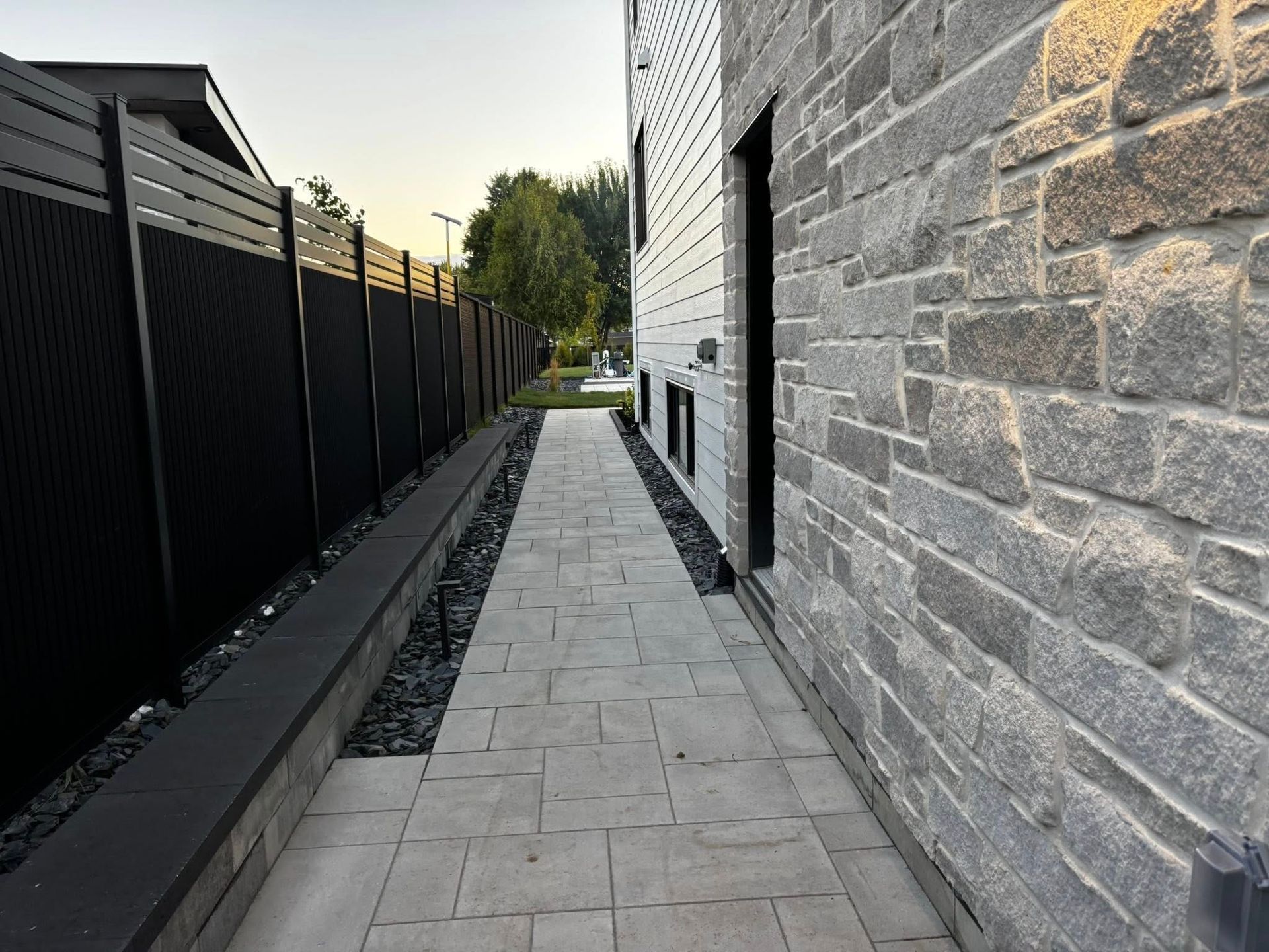 Trottoir entre une clôture sombre et un bâtiment revêtu de pierre.