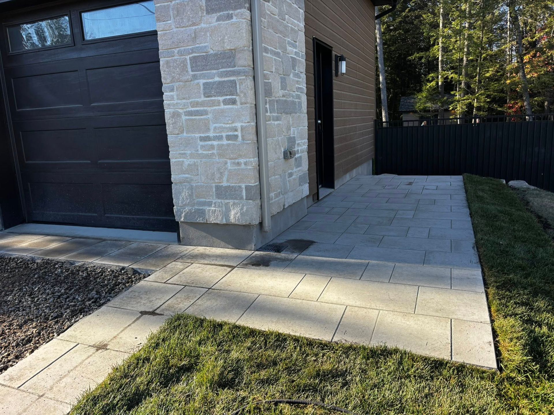 Un chemin pavé de dalles grises mène à un bâtiment avec une porte de garage noire et une façade en pierre.